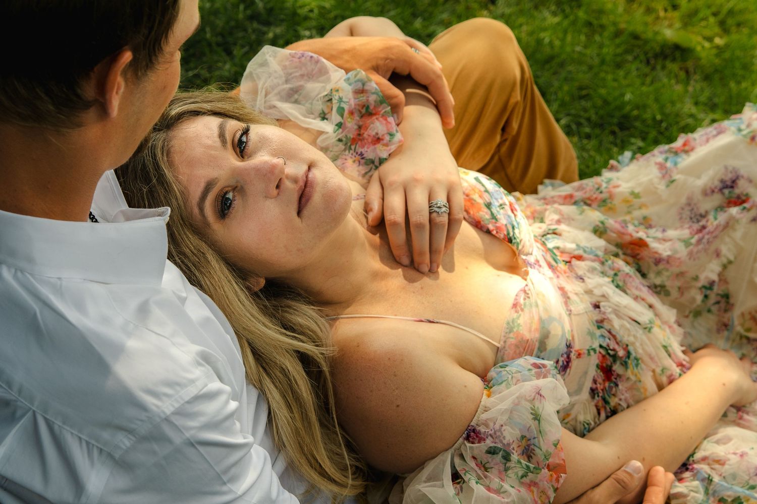 Couple relaxing outdoors on grass, woman in floral dress looking up at man in white shirt.