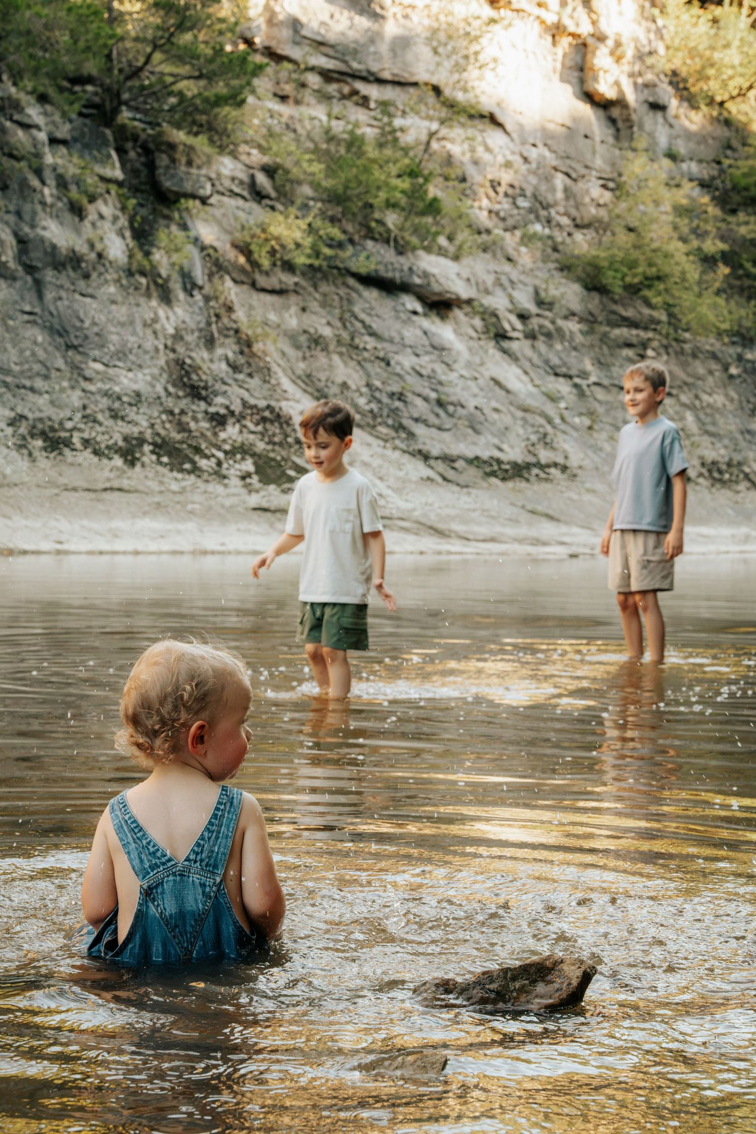 Three children playing in shallow river water near rocky cliffs on a sunny day.