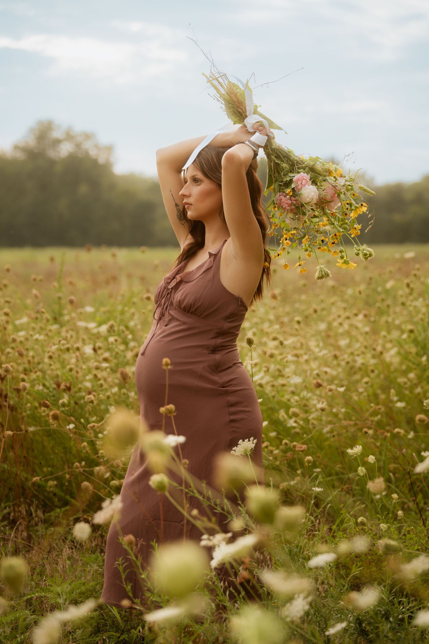 Pregnant woman in mauve dress holding wildflower bouquet overhead in blooming meadow during soft natural light.