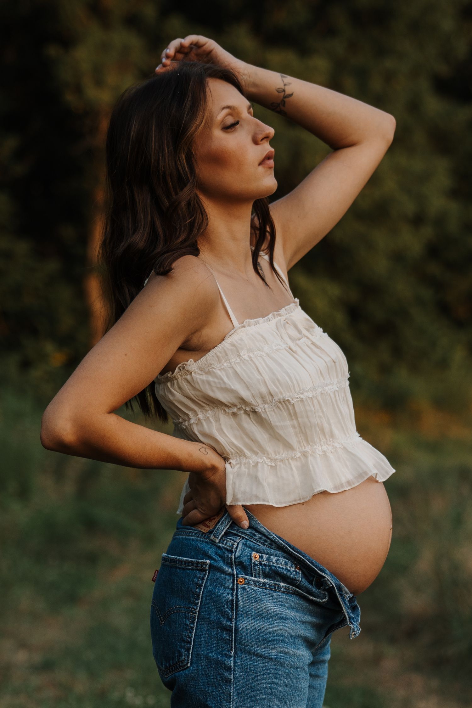 Pregnant woman in white crop top and unbuttoned jeans posing outdoors with hand on hip and arm raised.