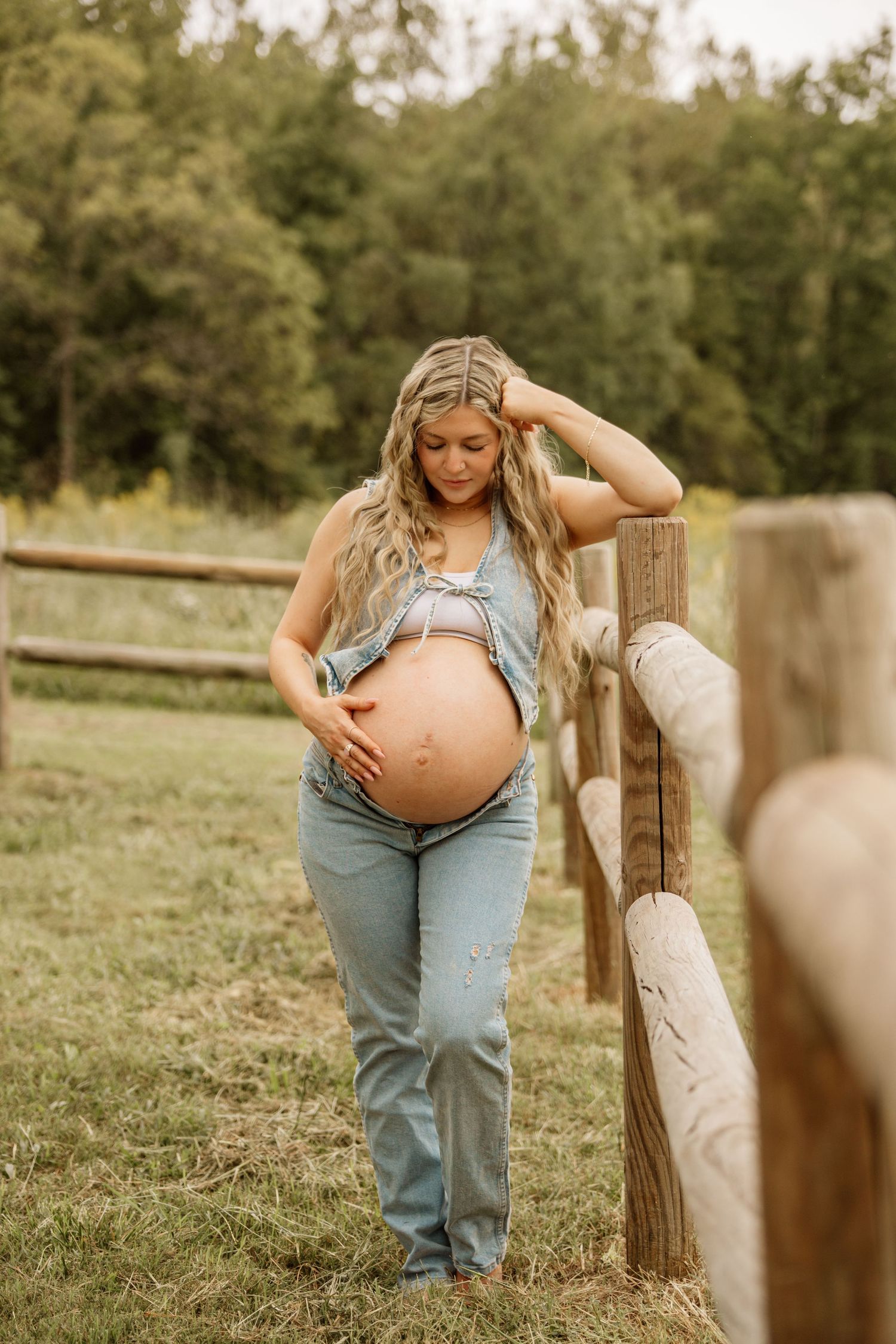 Pregnant woman in denim overalls leans against a wooden fence in a lush green outdoor setting.