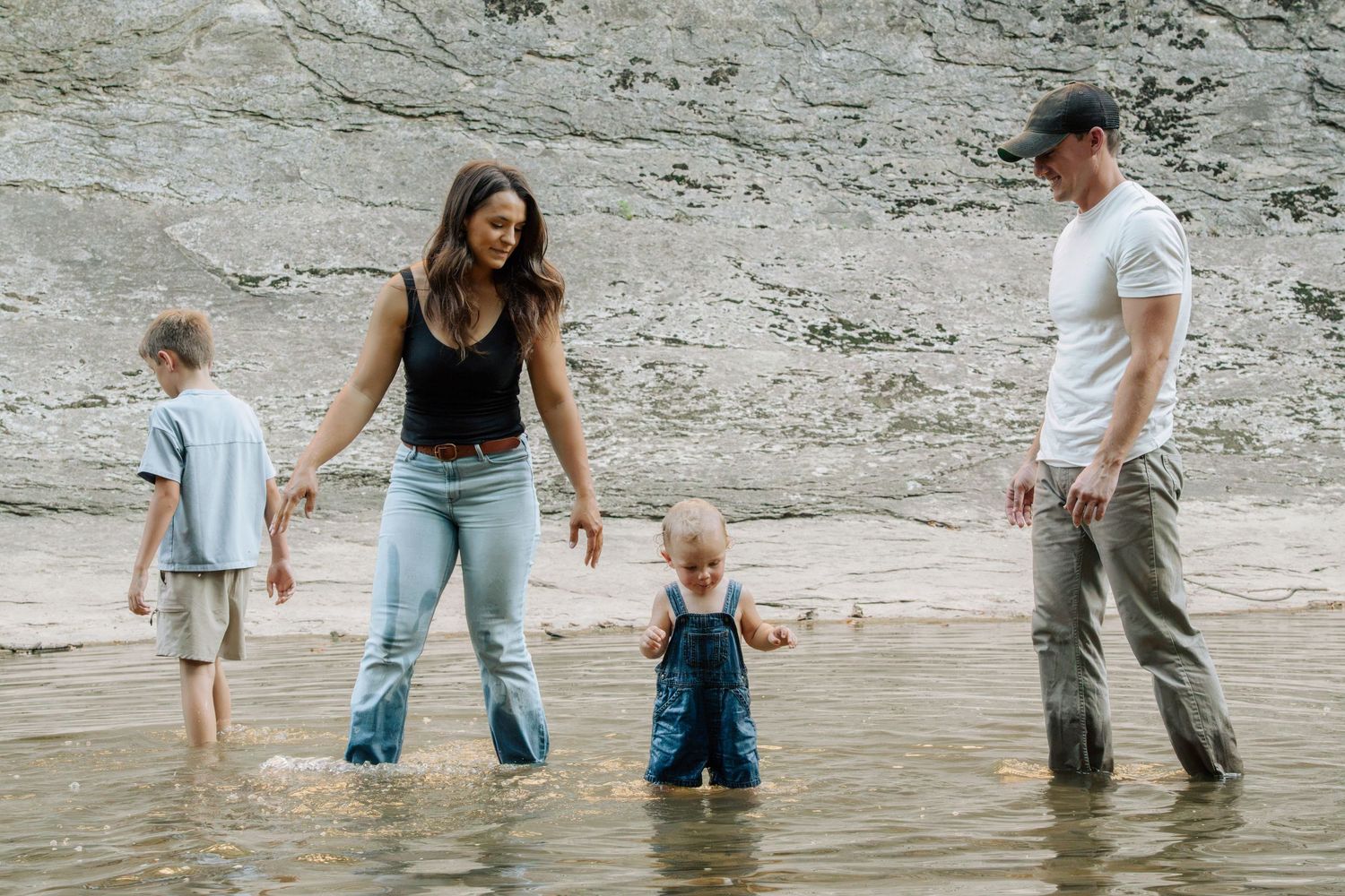 Family wading in shallow water near rocky cliffs, parents watching young children play at the beach.