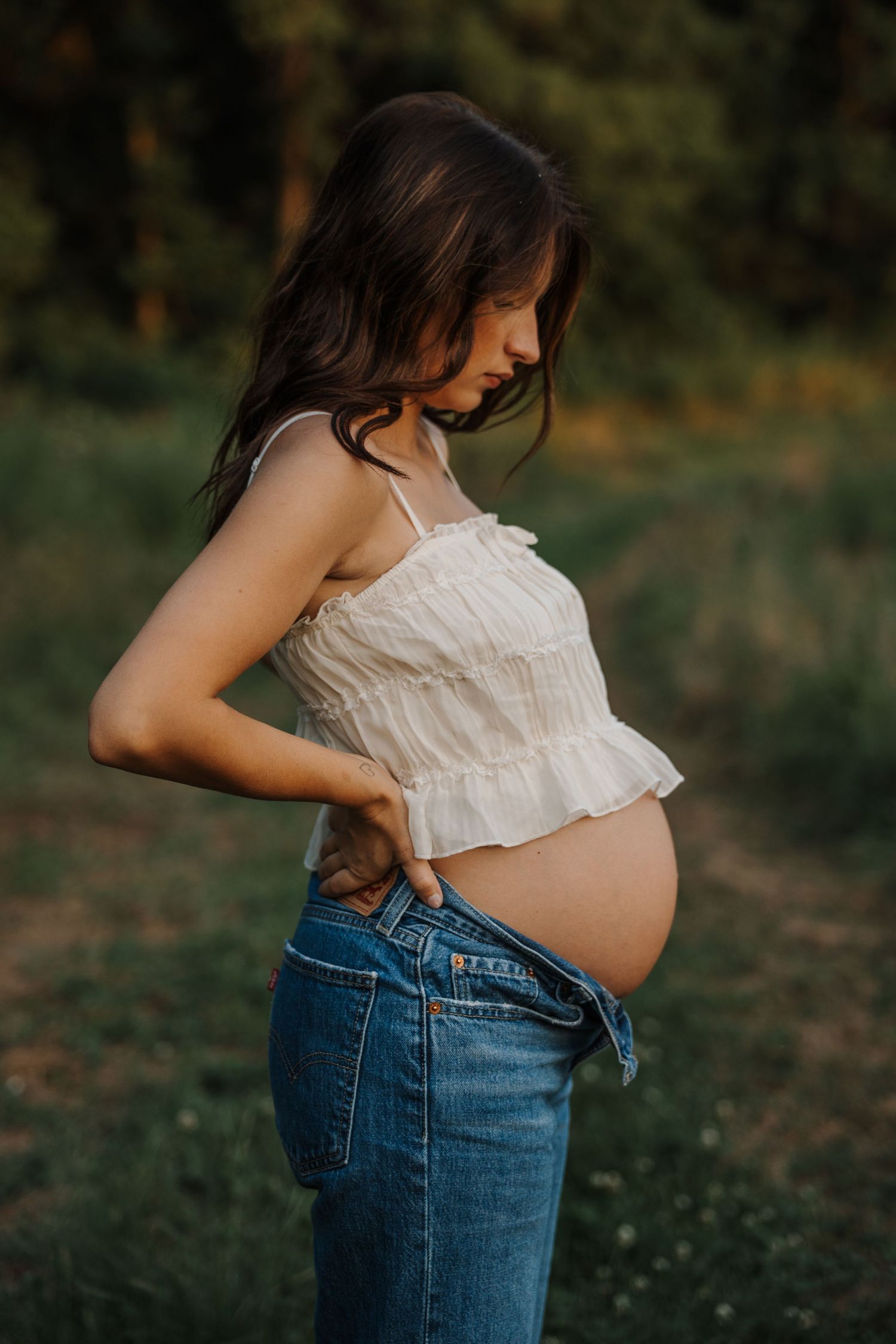 Pregnant woman in white crop top and jeans cradles bare baby bump in golden outdoor light.