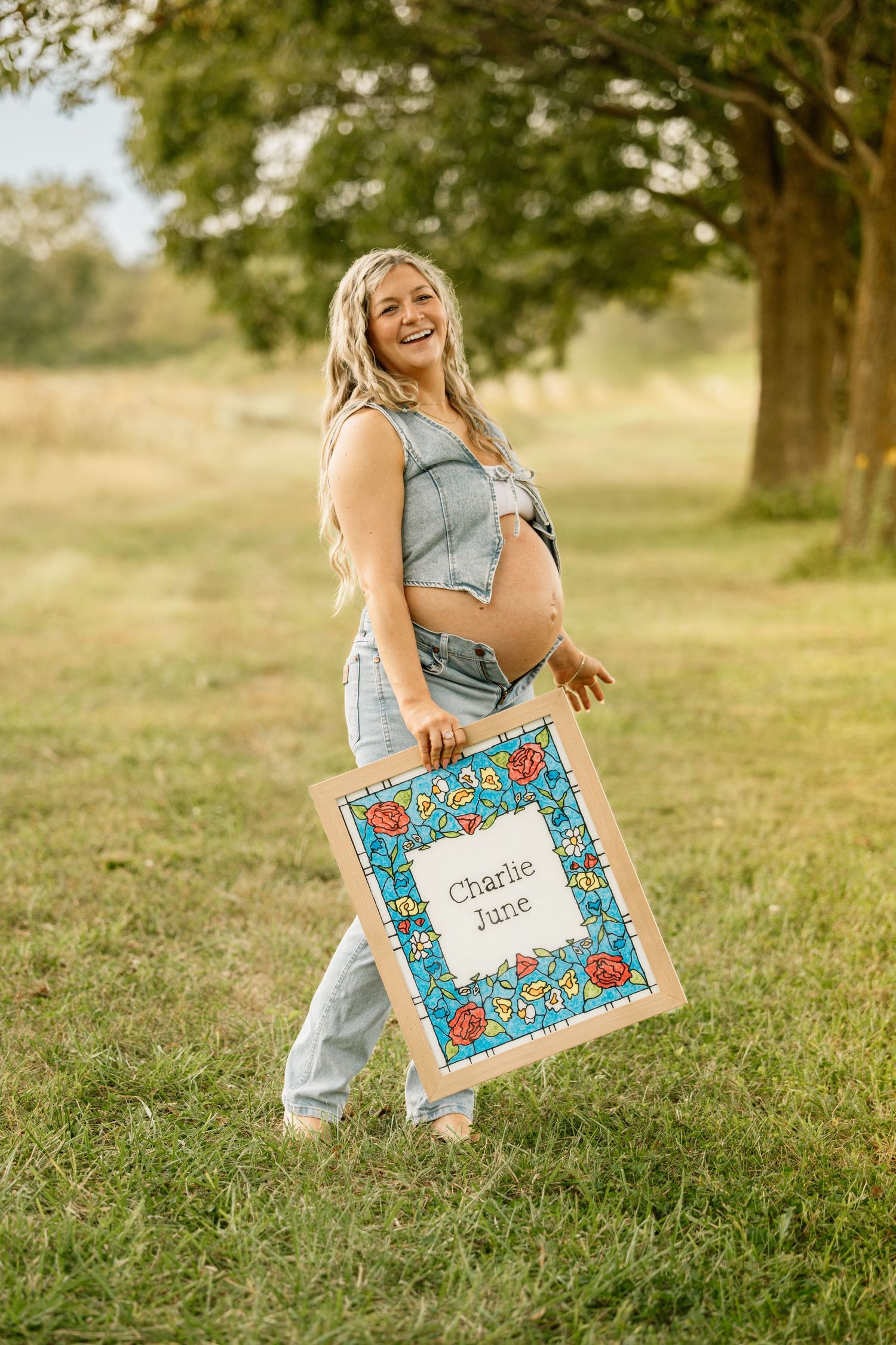 Smiling pregnant woman holding a floral framed sign reading Charlie June outdoors in a sunny green field.
