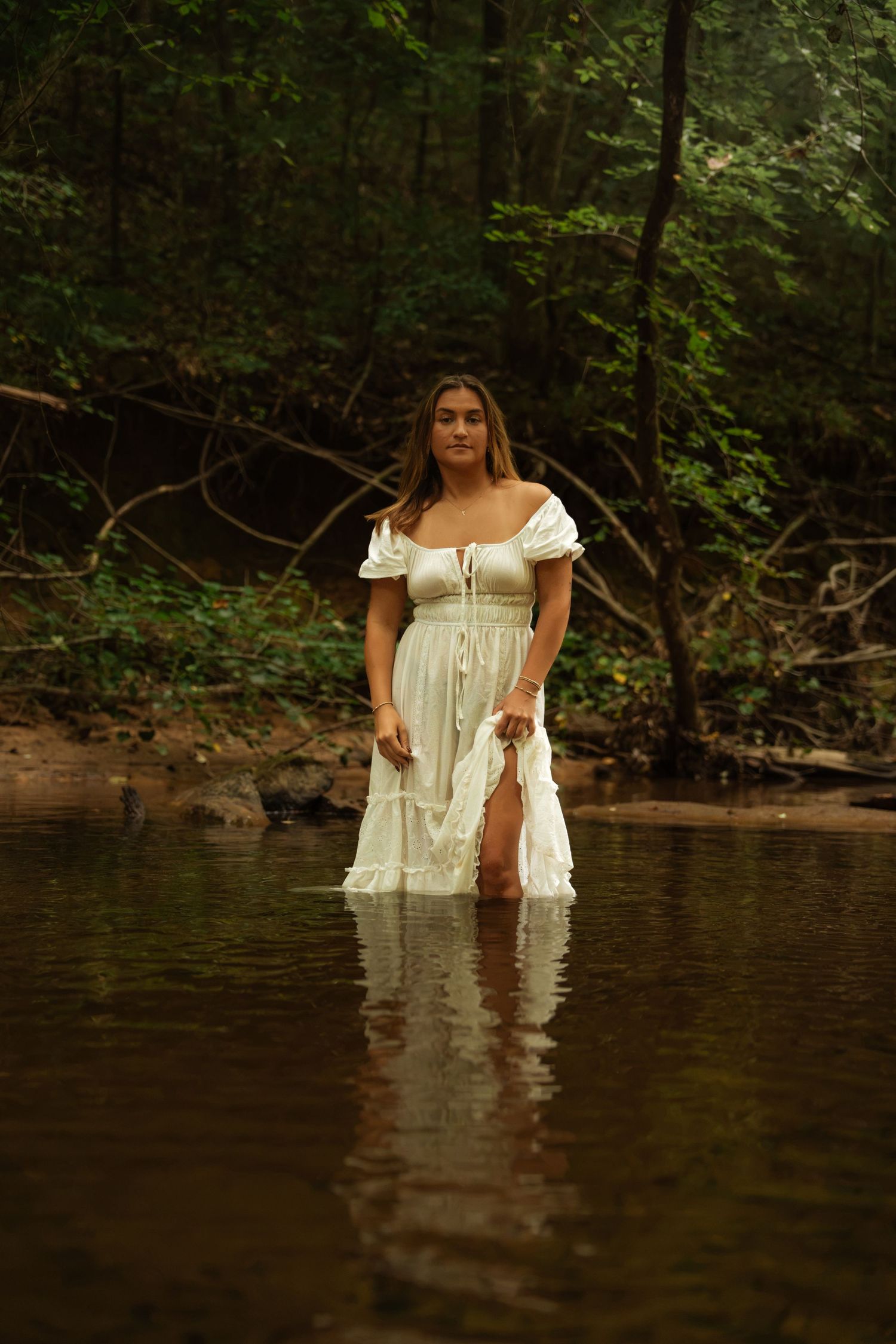 Woman in white off-shoulder dress standing in a forest creek surrounded by lush green trees.