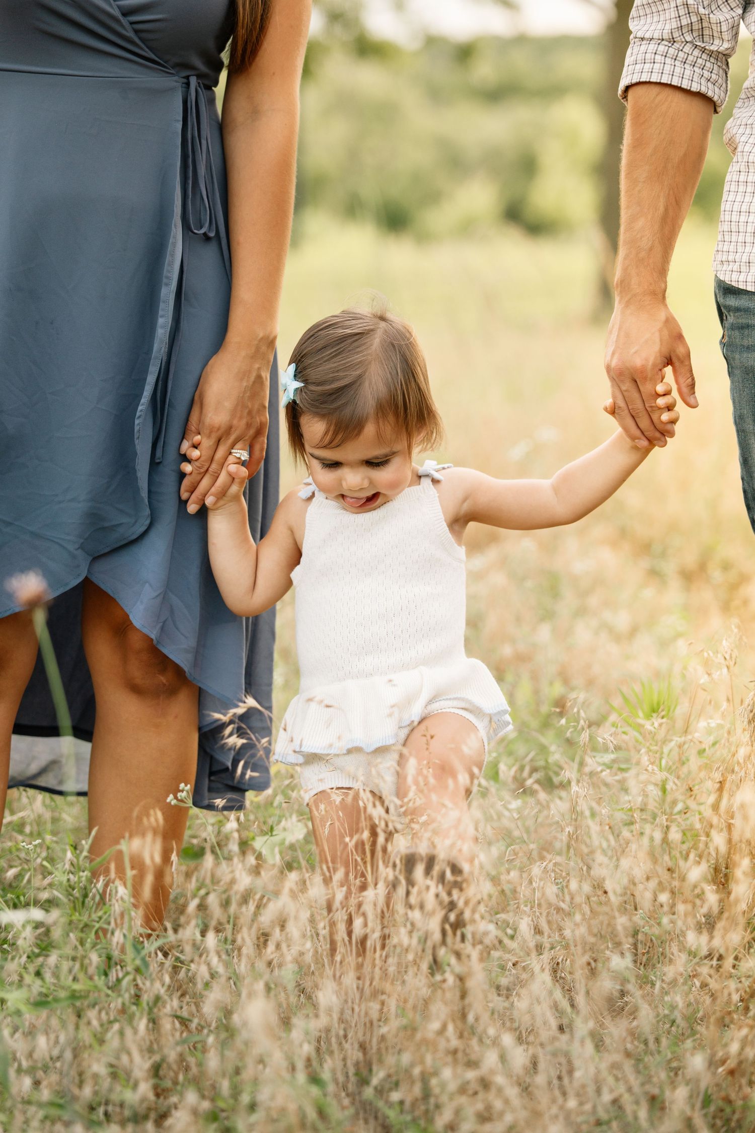 A toddler girl in white dress walks through a sunny meadow holding hands with her parents.