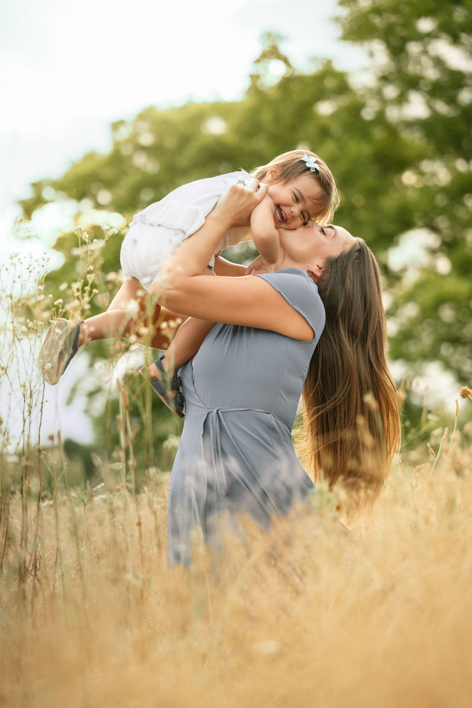 Joyful mother in blue dress lifting laughing toddler girl in golden summer field.