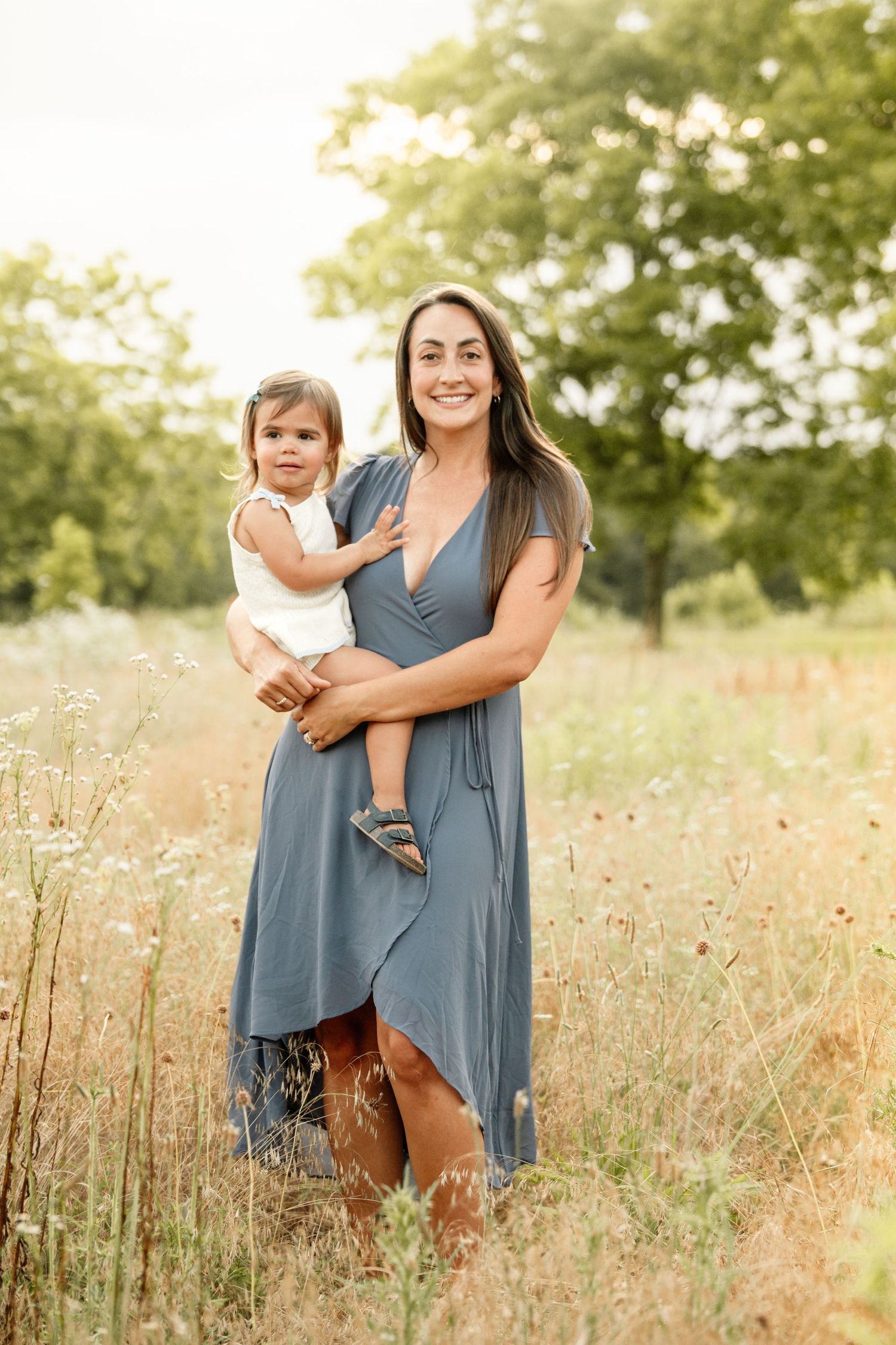 Smiling mother in blue dress holding her baby girl while standing in a wildflower meadow.