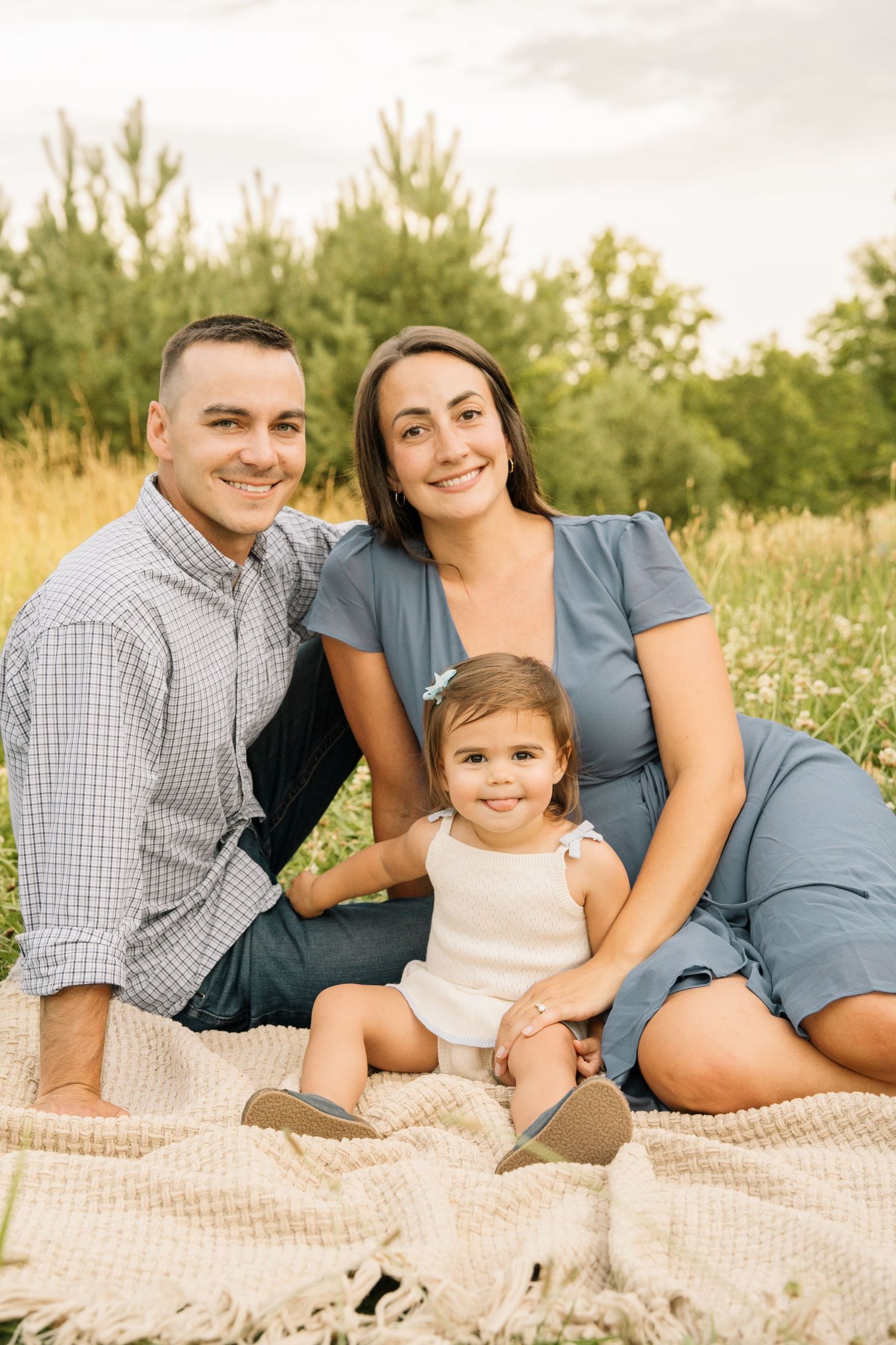 Family of three sitting on blanket in field, parents smiling with toddler daughter in white dress.