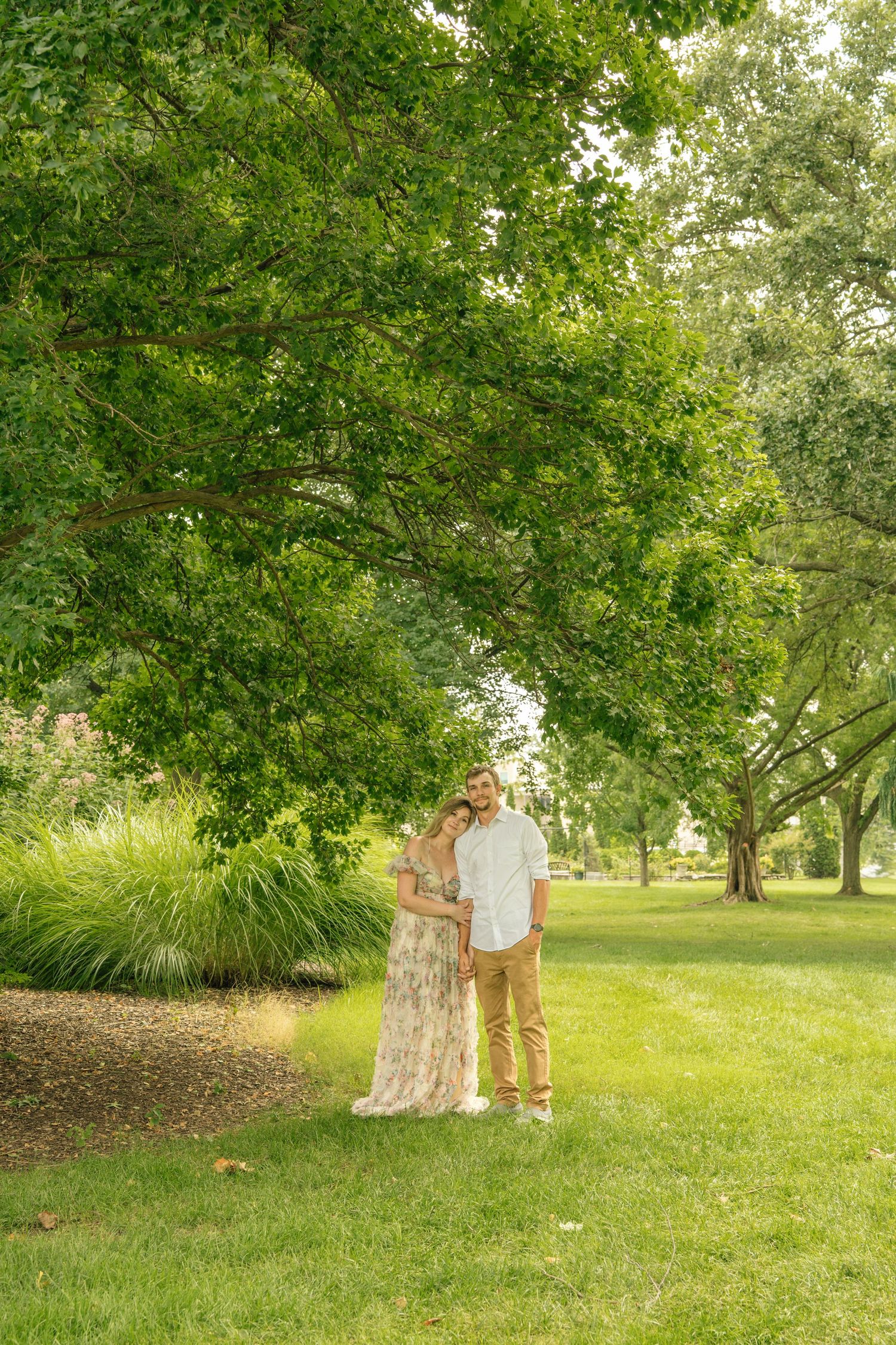 Couple standing together under a large green tree in a serene park setting.