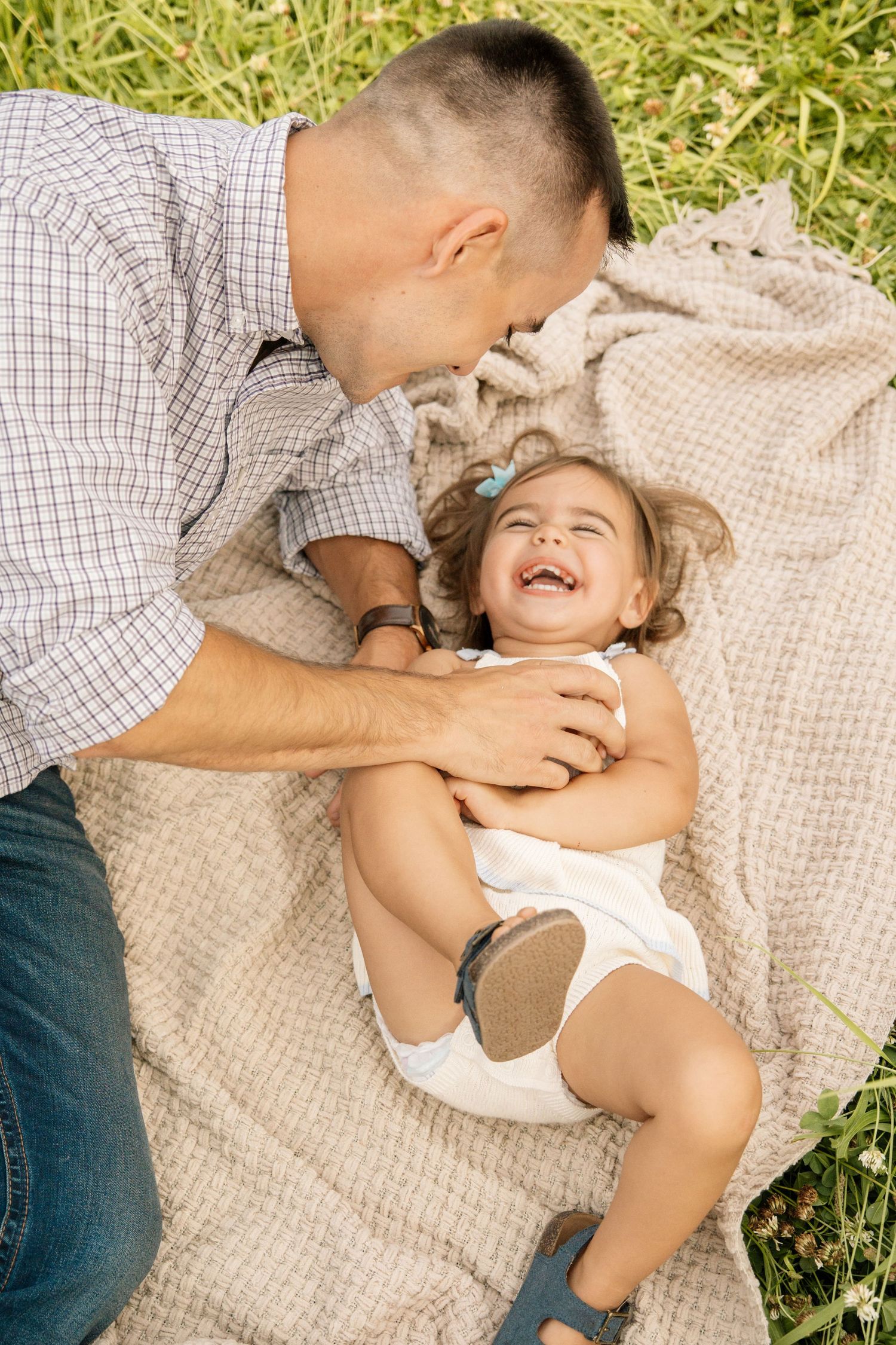 Father tickling laughing toddler girl on blanket in green grass field during outdoor family picnic.