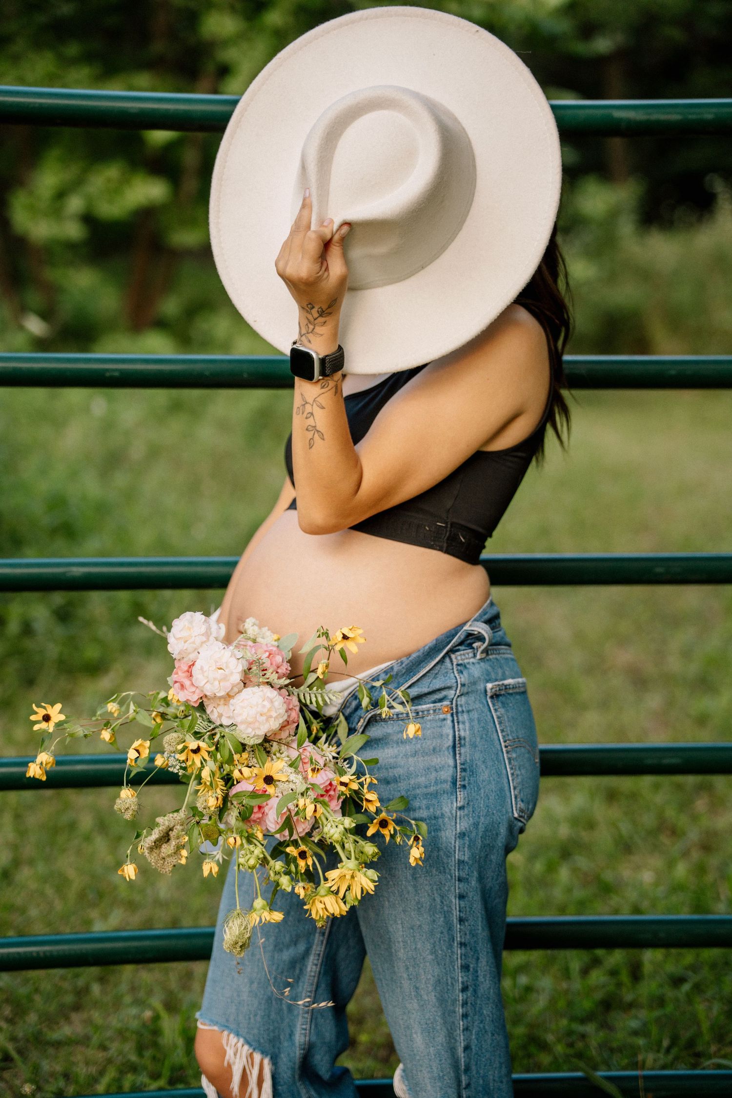 Pregnant woman in jeans holding white hat over face with wildflower bouquet near green metal fence outdoors.