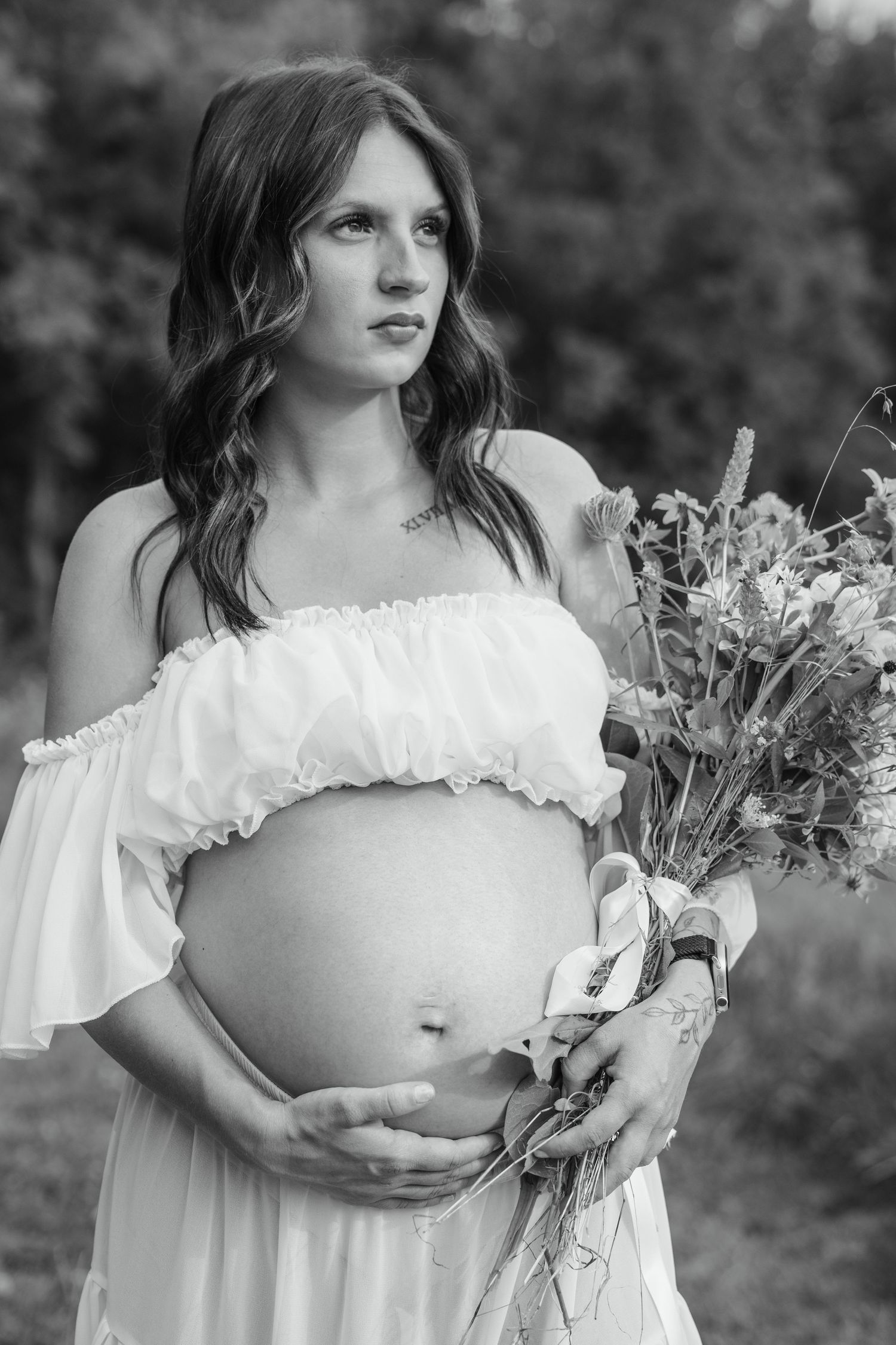 Black and white portrait of a pregnant woman in white dress holding wildflowers outdoors.
