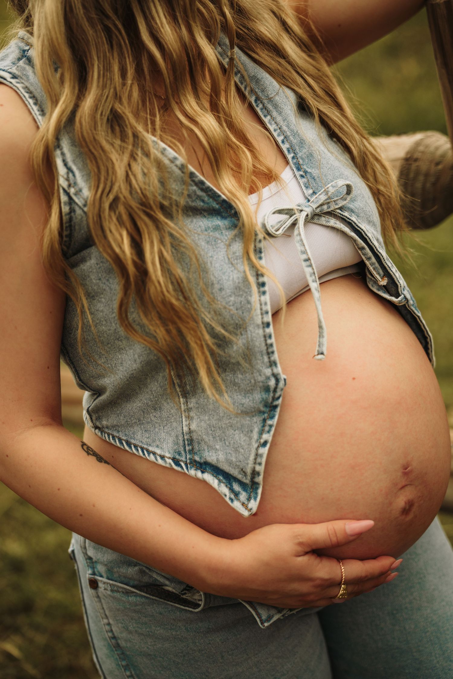 Close-up of pregnant woman in denim vest cradling bare baby bump with gold rings on fingers.