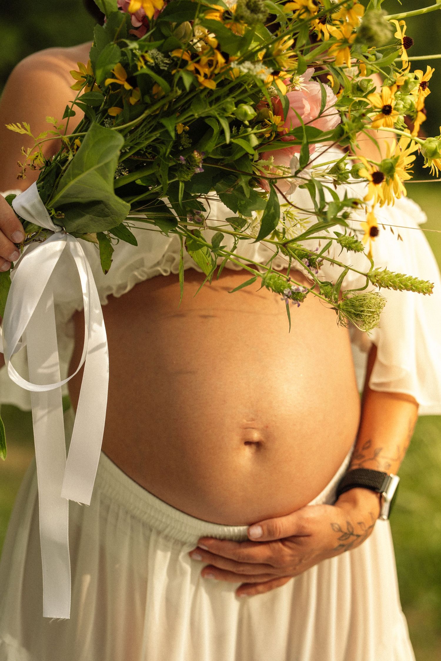 Pregnant woman in white dress holding wildflower bouquet with ribbon, cradling bare baby bump outdoors.