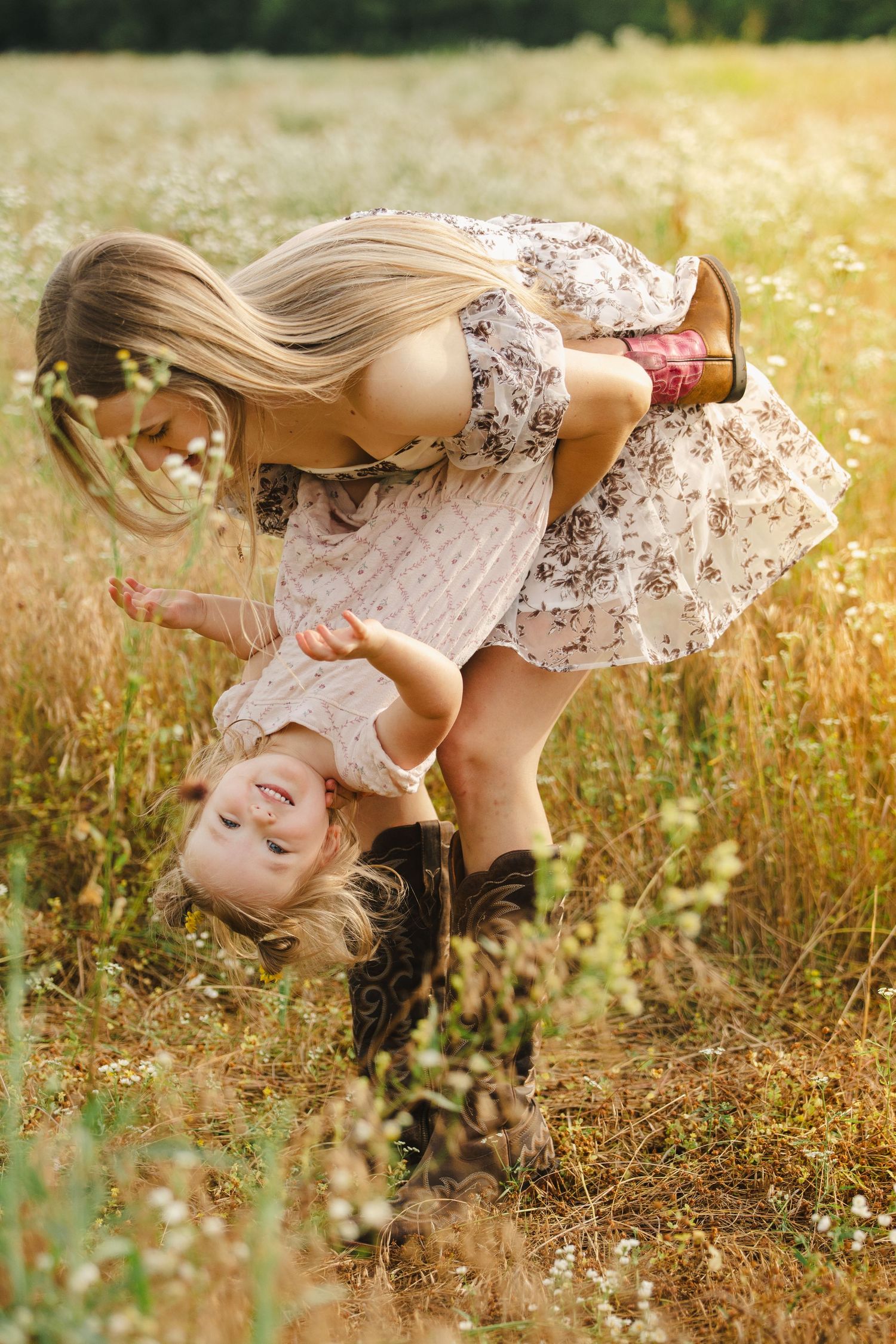 Mother playfully holding toddler upside down in a sunny wildflower meadow, both wearing floral dresses.