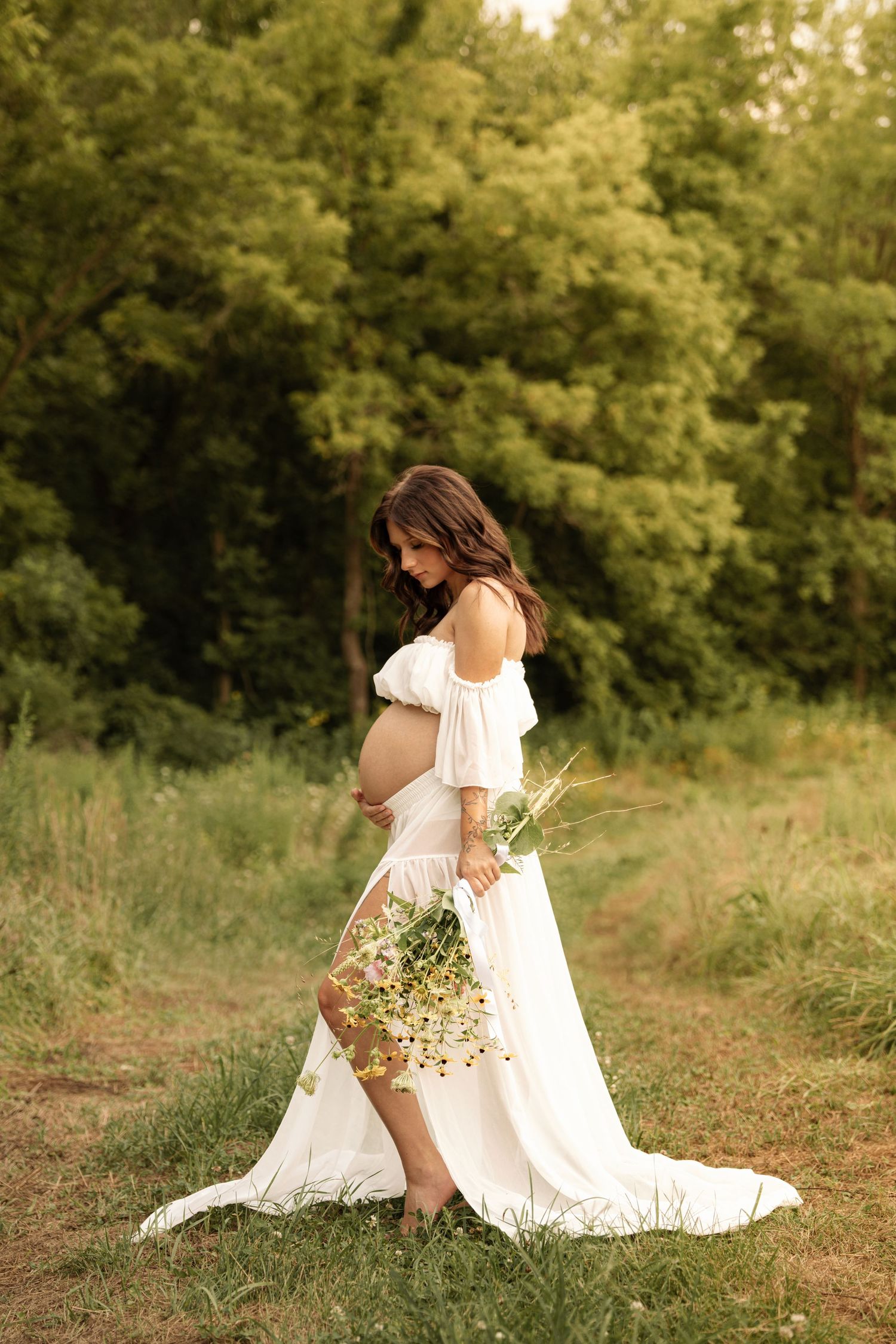 Pregnant woman in white off-shoulder dress holding wildflowers, posing outdoors in a green nature setting.