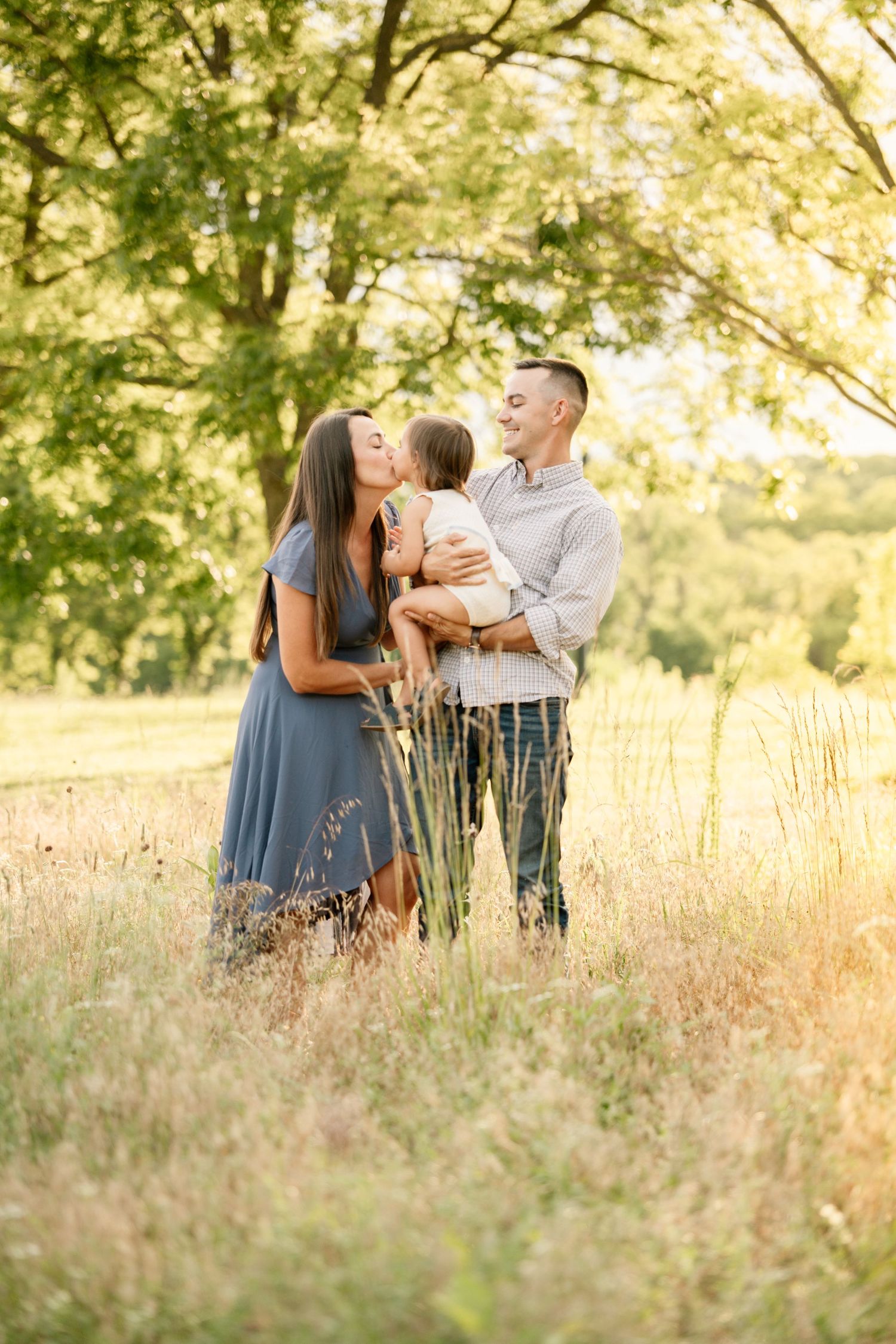 Happy family with parents kissing their toddler daughter in a golden sunlit meadow.