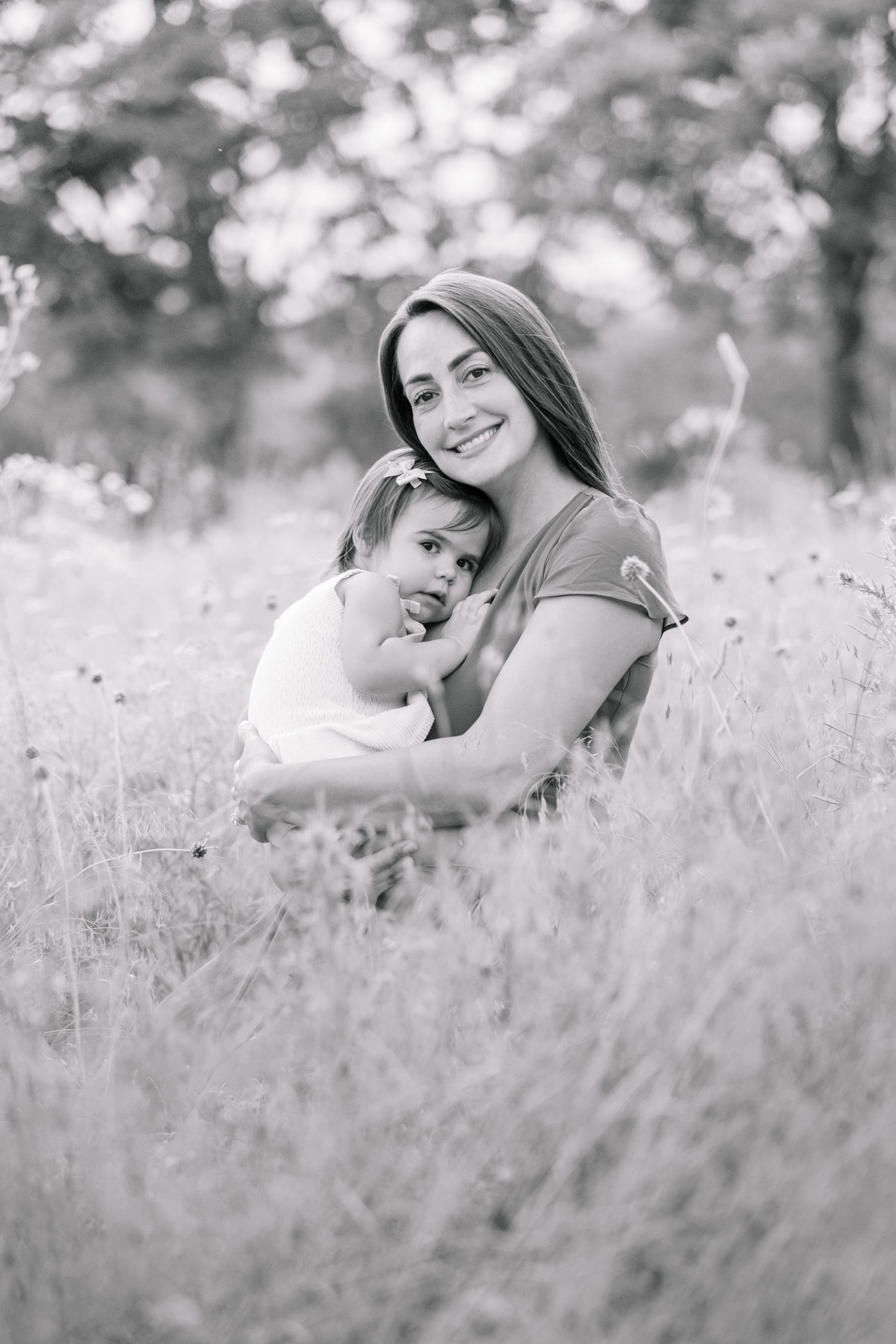 Black and white portrait of a smiling mother embracing her toddler daughter in an outdoor meadow.