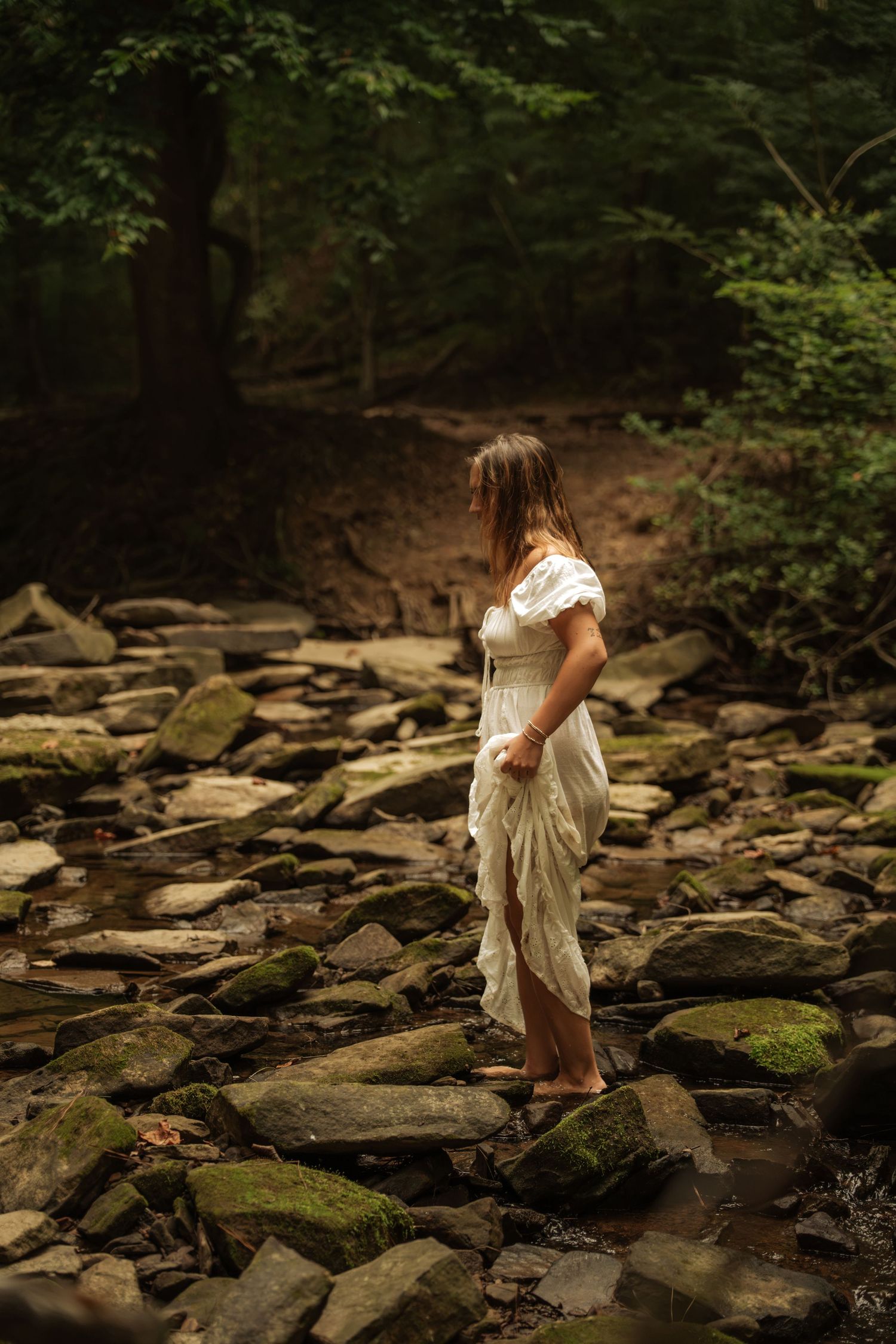 Woman in white dress standing barefoot on mossy rocks in a forest stream, surrounded by lush green trees.