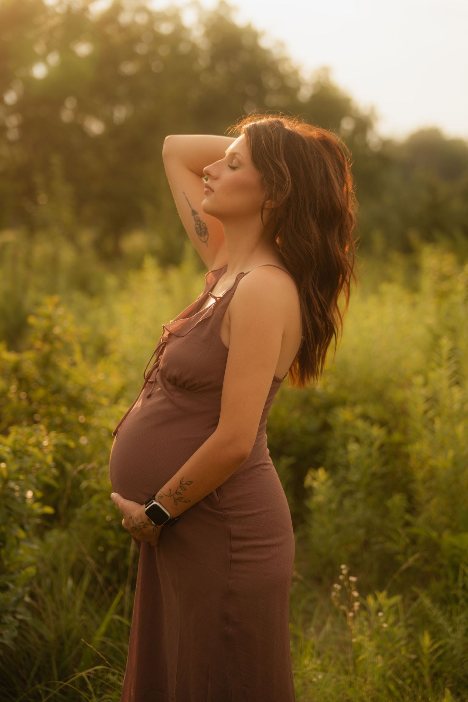 Pregnant woman in brown dress standing in green field during golden hour sunset, cradling baby bump with eyes closed.