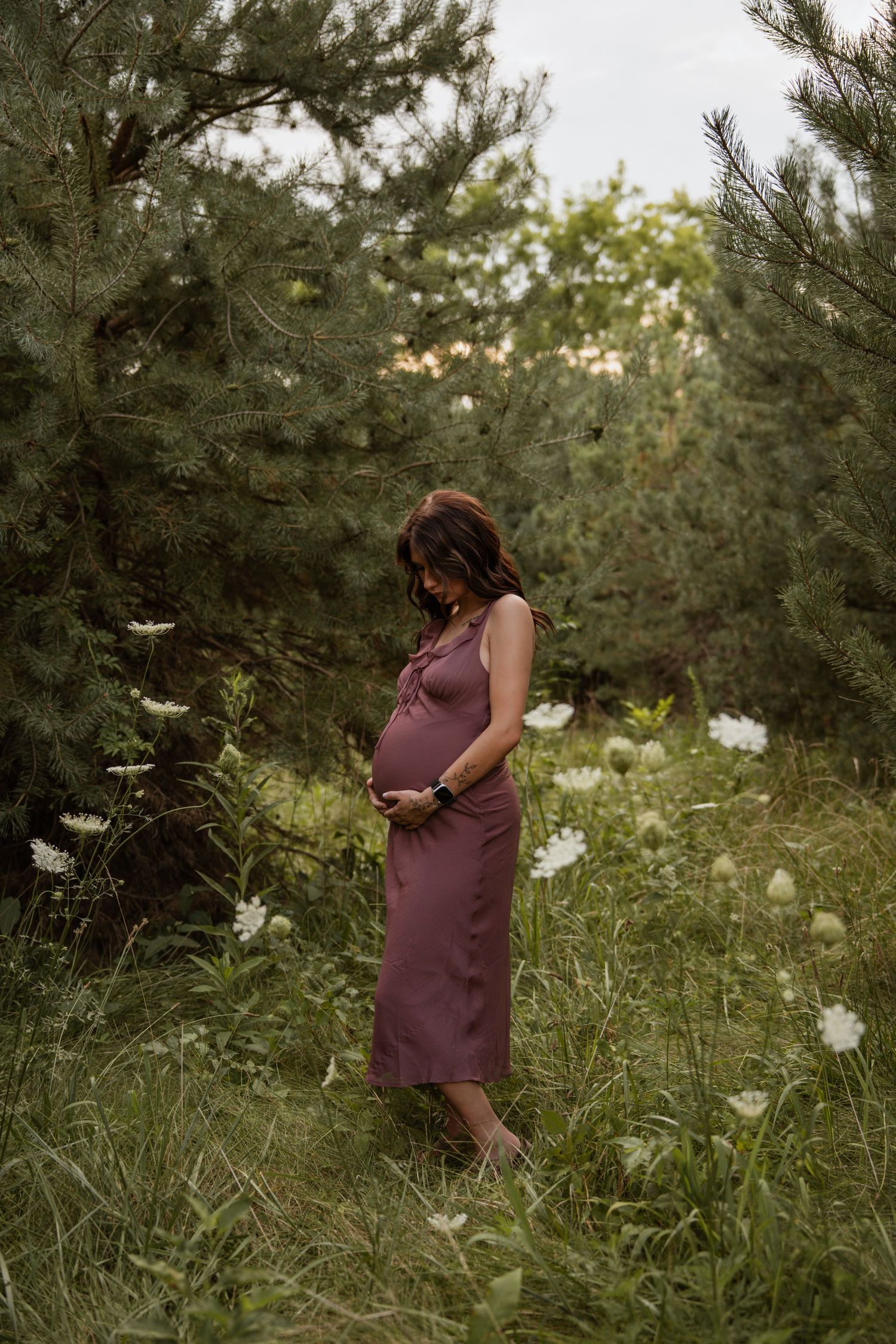 Pregnant woman in dusty rose dress cradles baby bump while standing among pine trees and wildflowers.