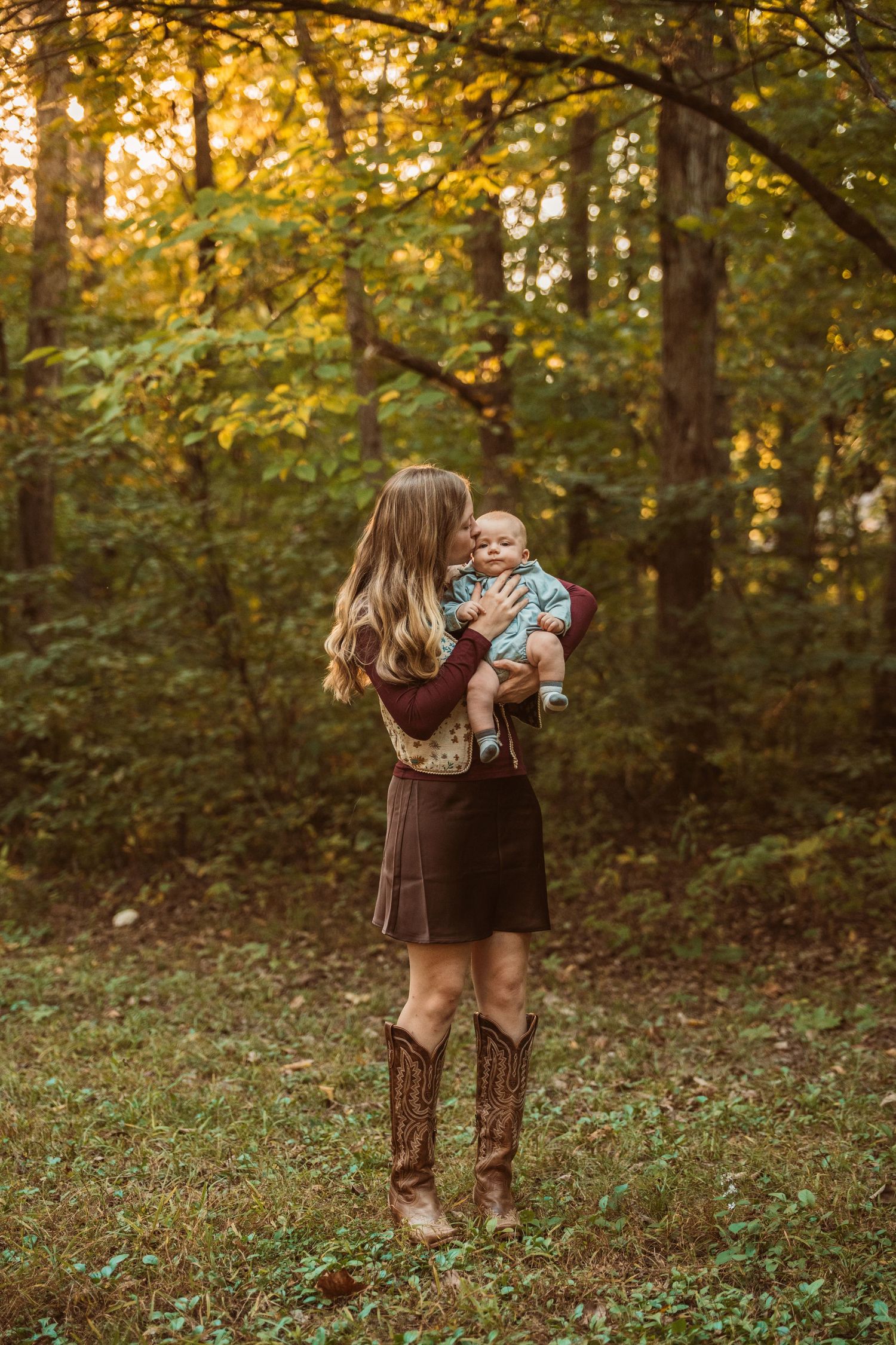A mother in cowboy boots holds her baby in a sunlit autumn forest clearing.