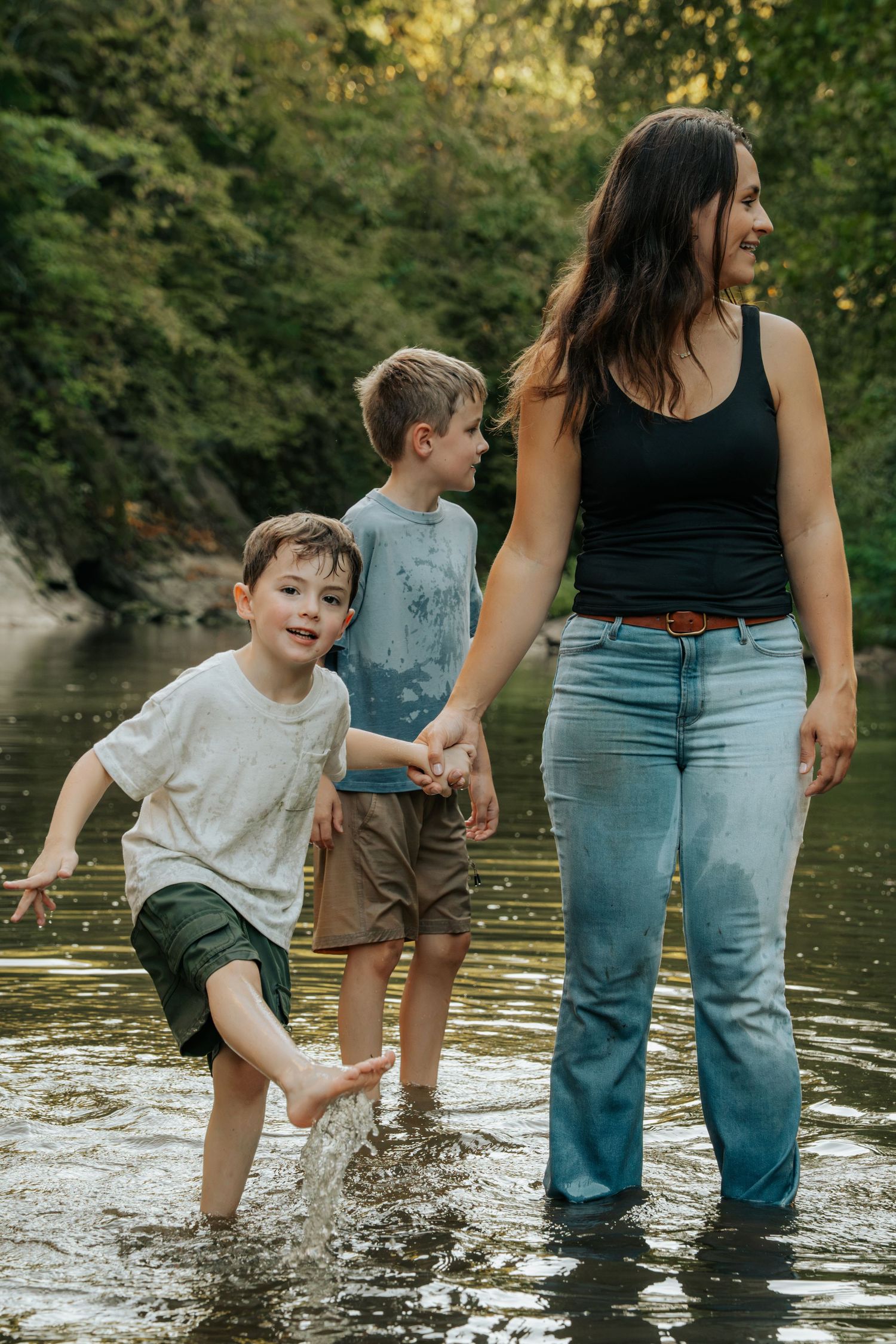 A mother and two young boys wade and splash in a shallow creek surrounded by lush green trees.