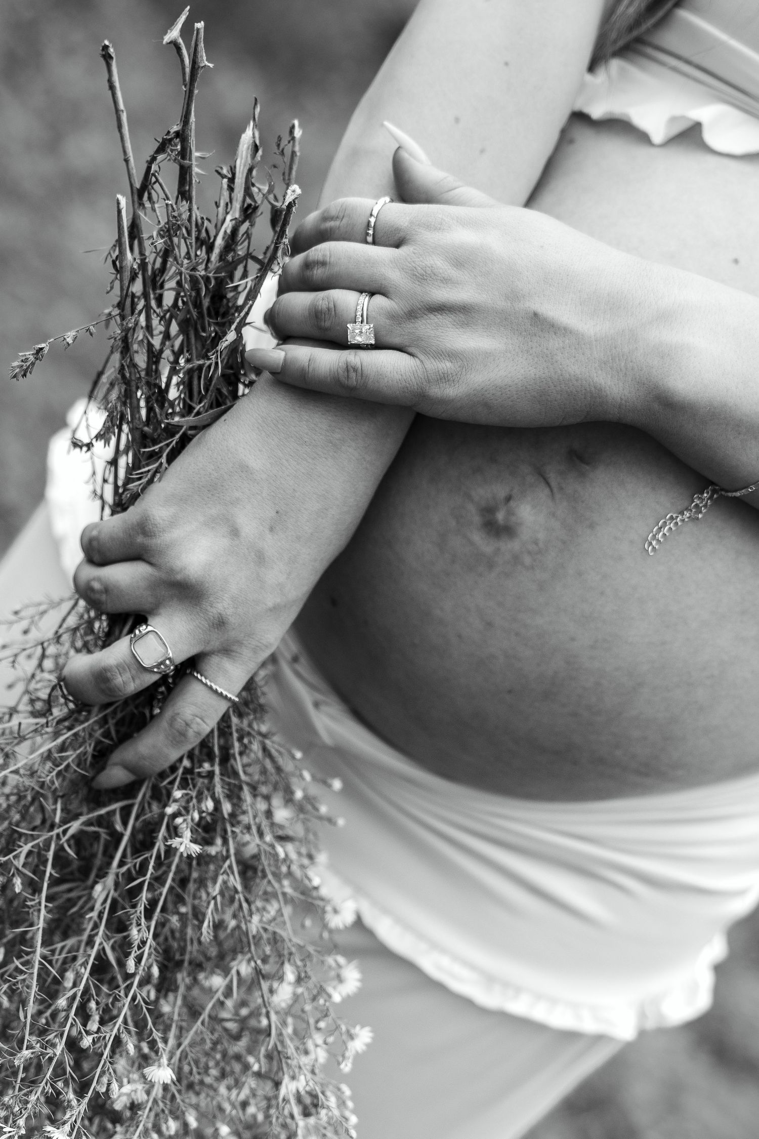 Black and white photo of pregnant woman cradling baby bump while holding wildflowers, wearing rings and delicate bracelet.