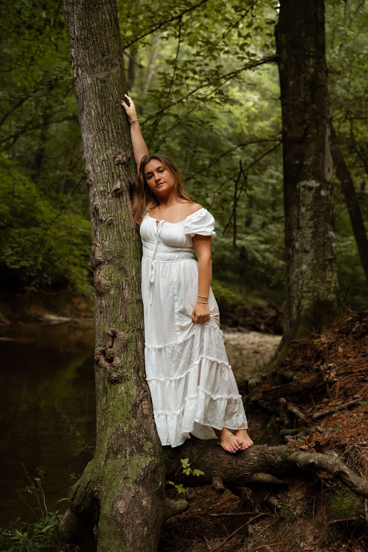 Barefoot woman in white dress leaning against a tree in a lush green forest.