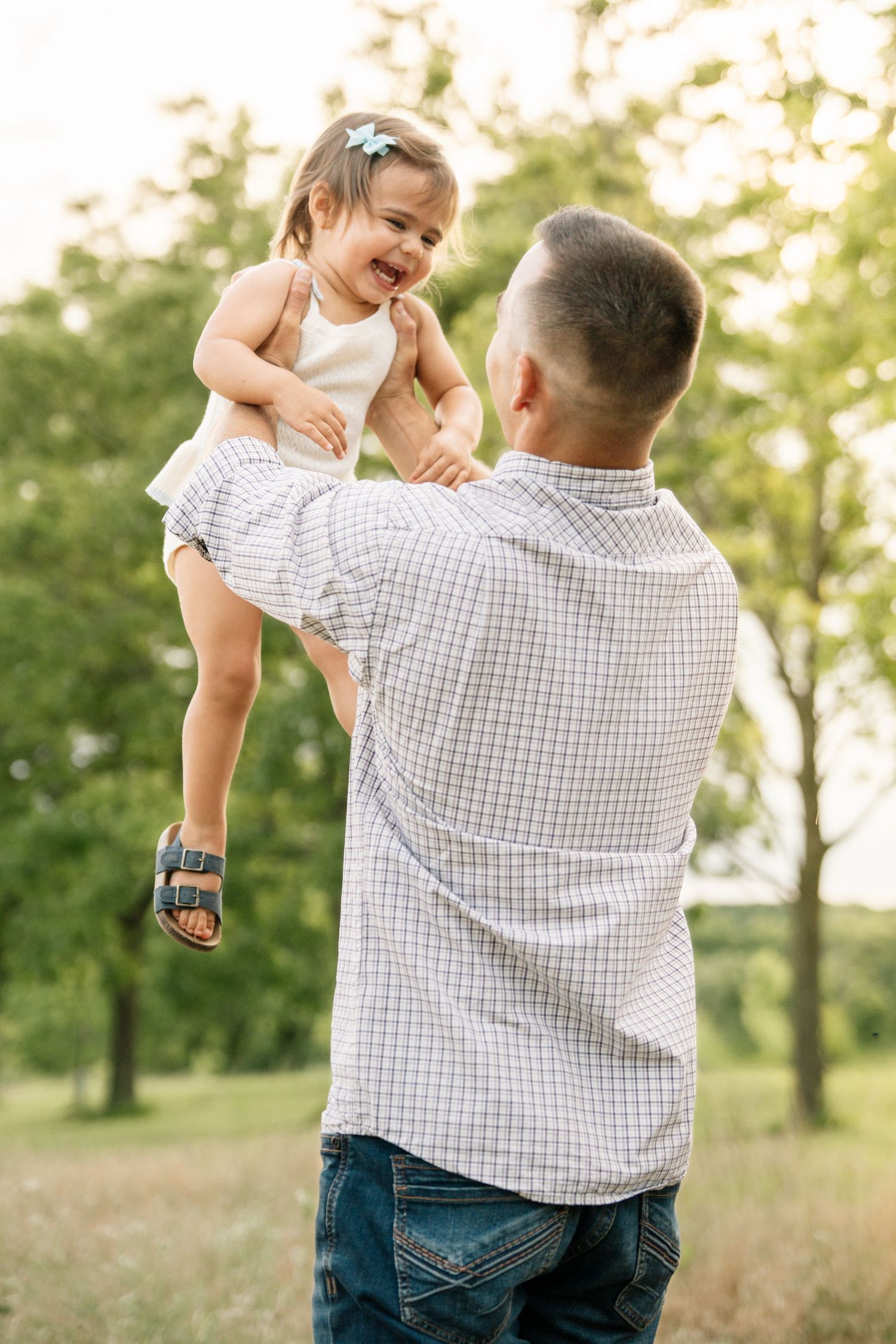 Father lifting his laughing toddler daughter in the air outdoors in a sunny green park.