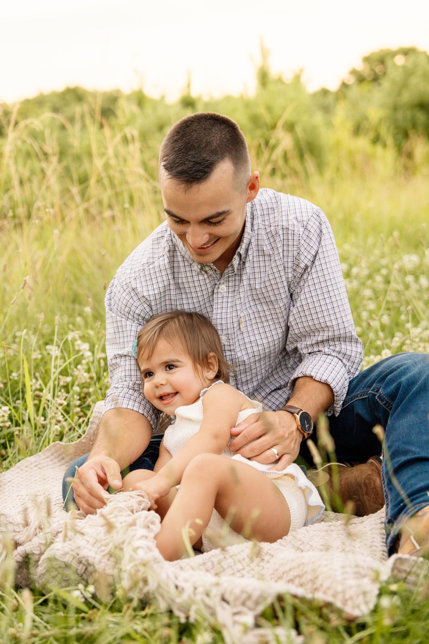 A laughing father sits on a blanket in a green field playfully holding his smiling toddler daughter.