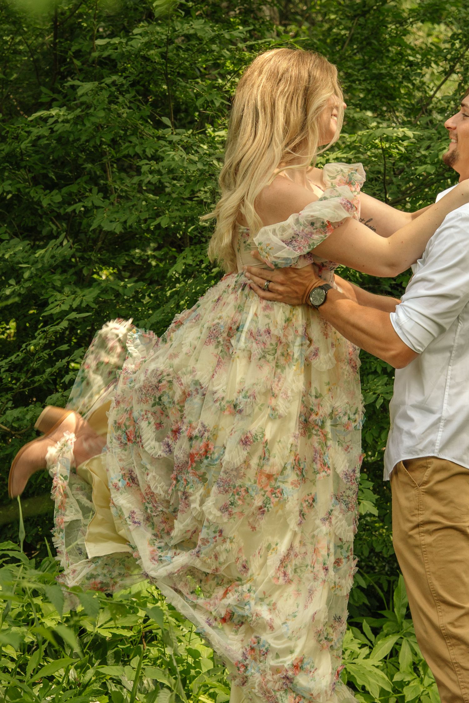 Man lifting woman in flowing floral dress outdoors surrounded by lush green foliage.