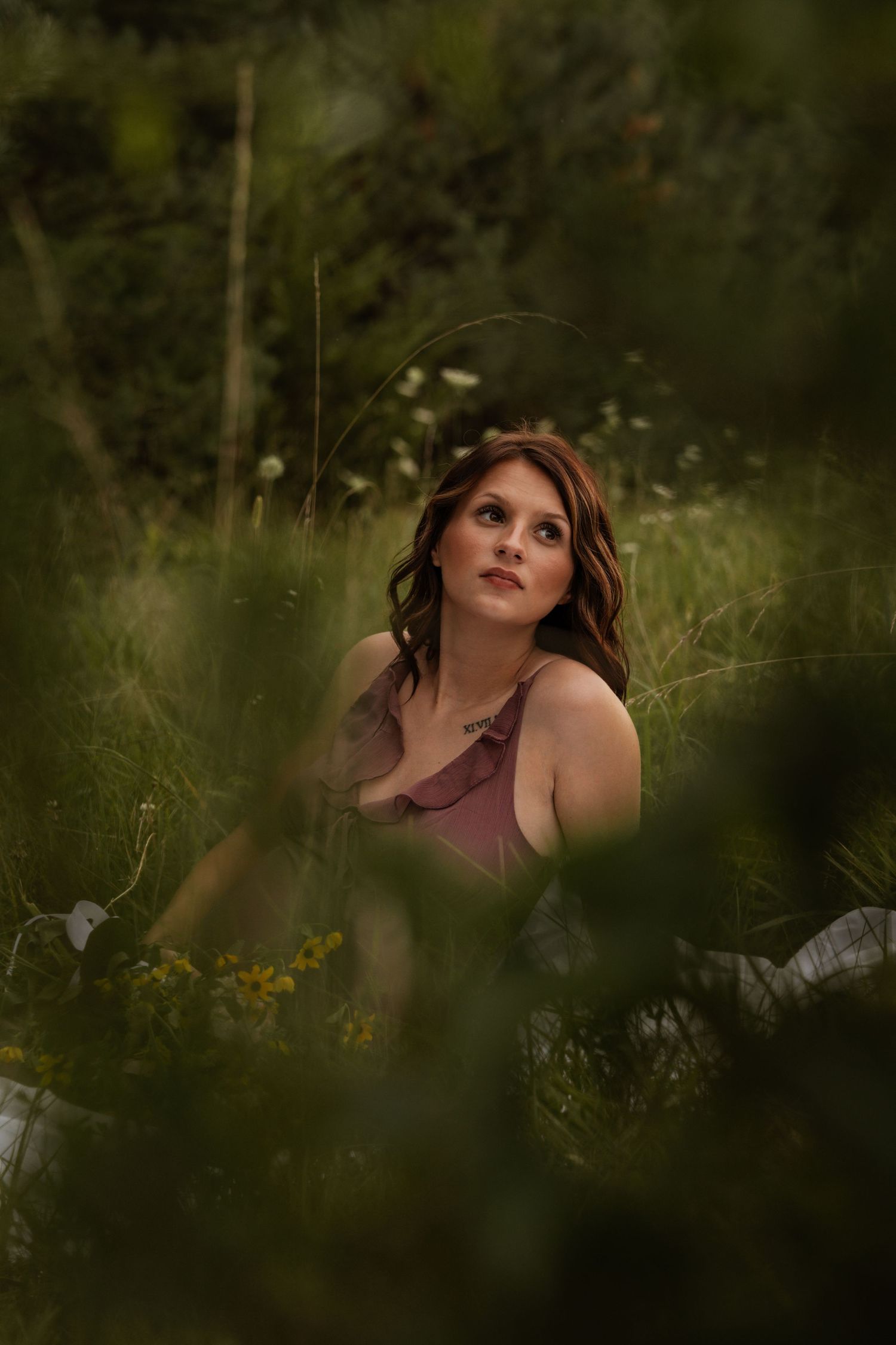 Pregnant woman in mauve dress sits among tall green grass and wildflowers gazing upward dreamily.