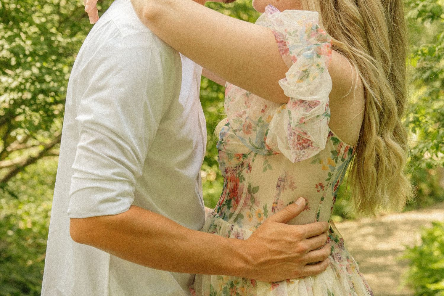 Couple embracing outdoors, woman in floral dress with long blonde hair, man in white shirt, surrounded by green trees.