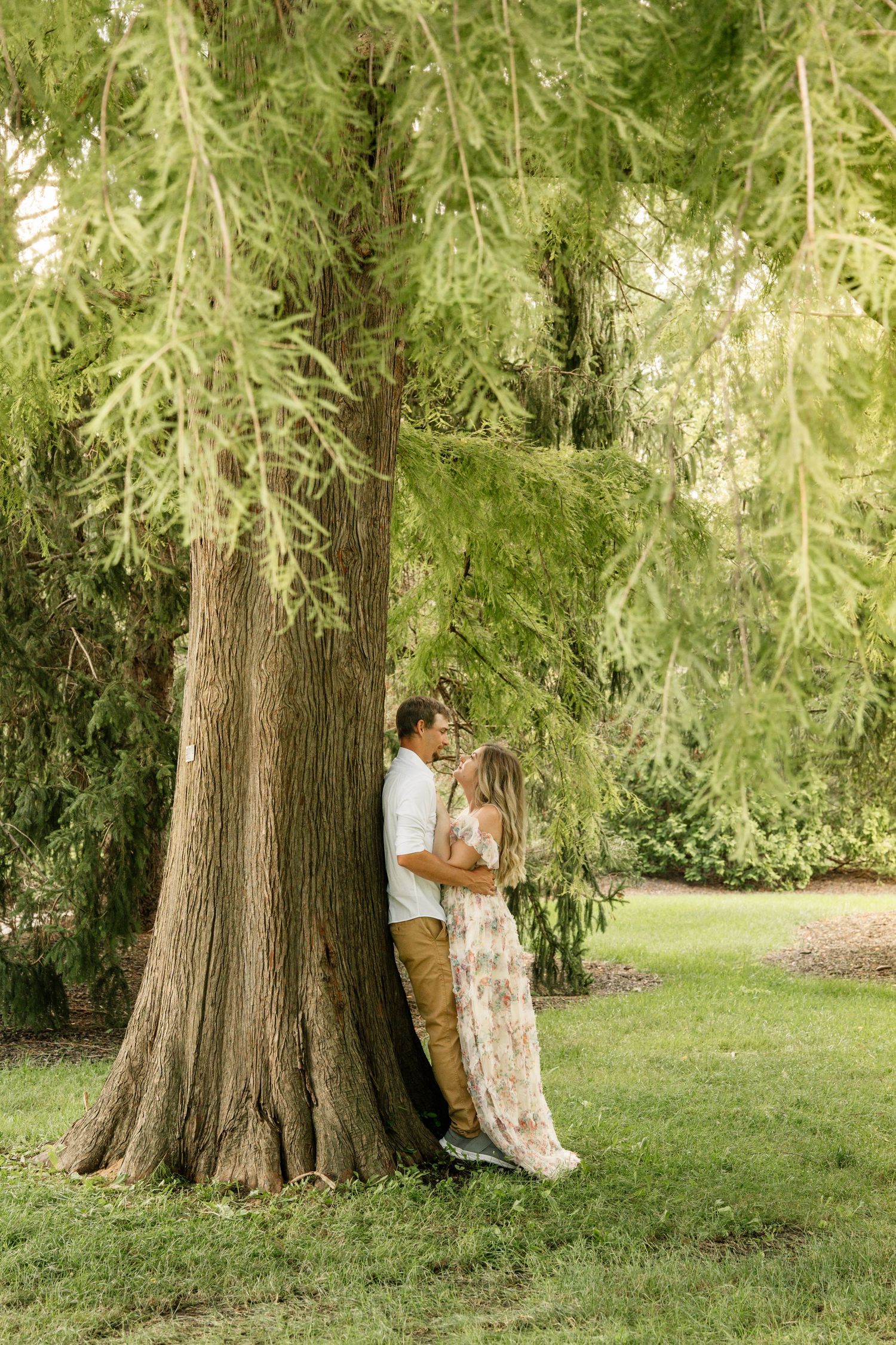 Couple sharing a romantic kiss while leaning against a large tree in a lush green park setting.