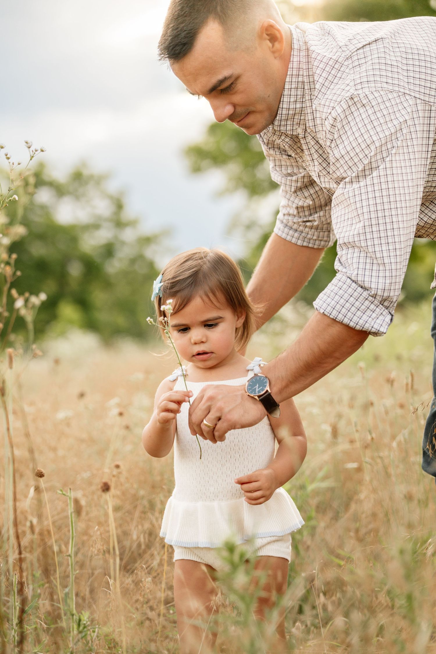 Father helping toddler daughter walk through golden wildflower field outdoors on a warm sunny day.