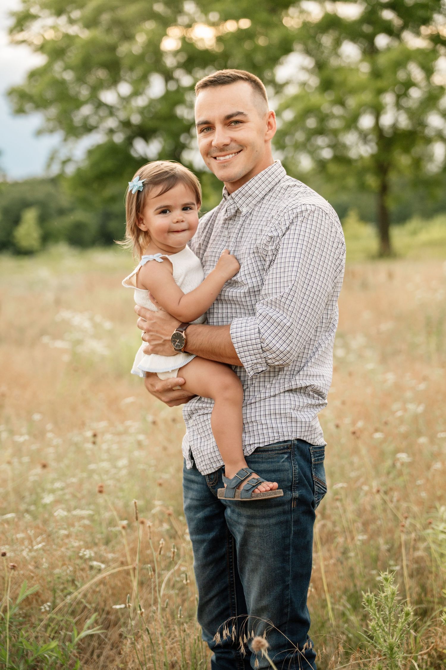 Smiling father holds his toddler daughter in a sunny wildflower field.