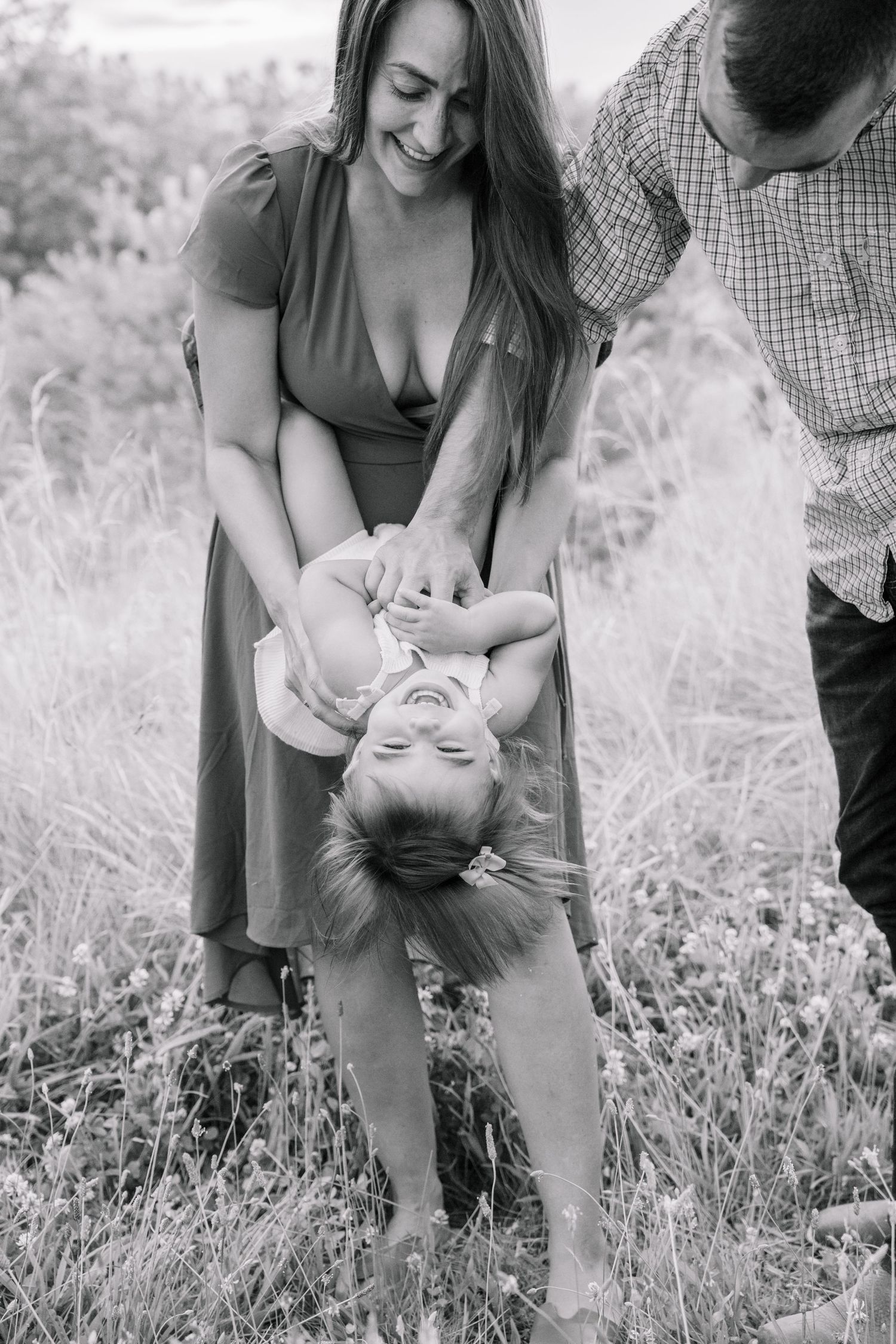 Black and white photo of laughing parents playfully holding toddler upside down in a grassy meadow.