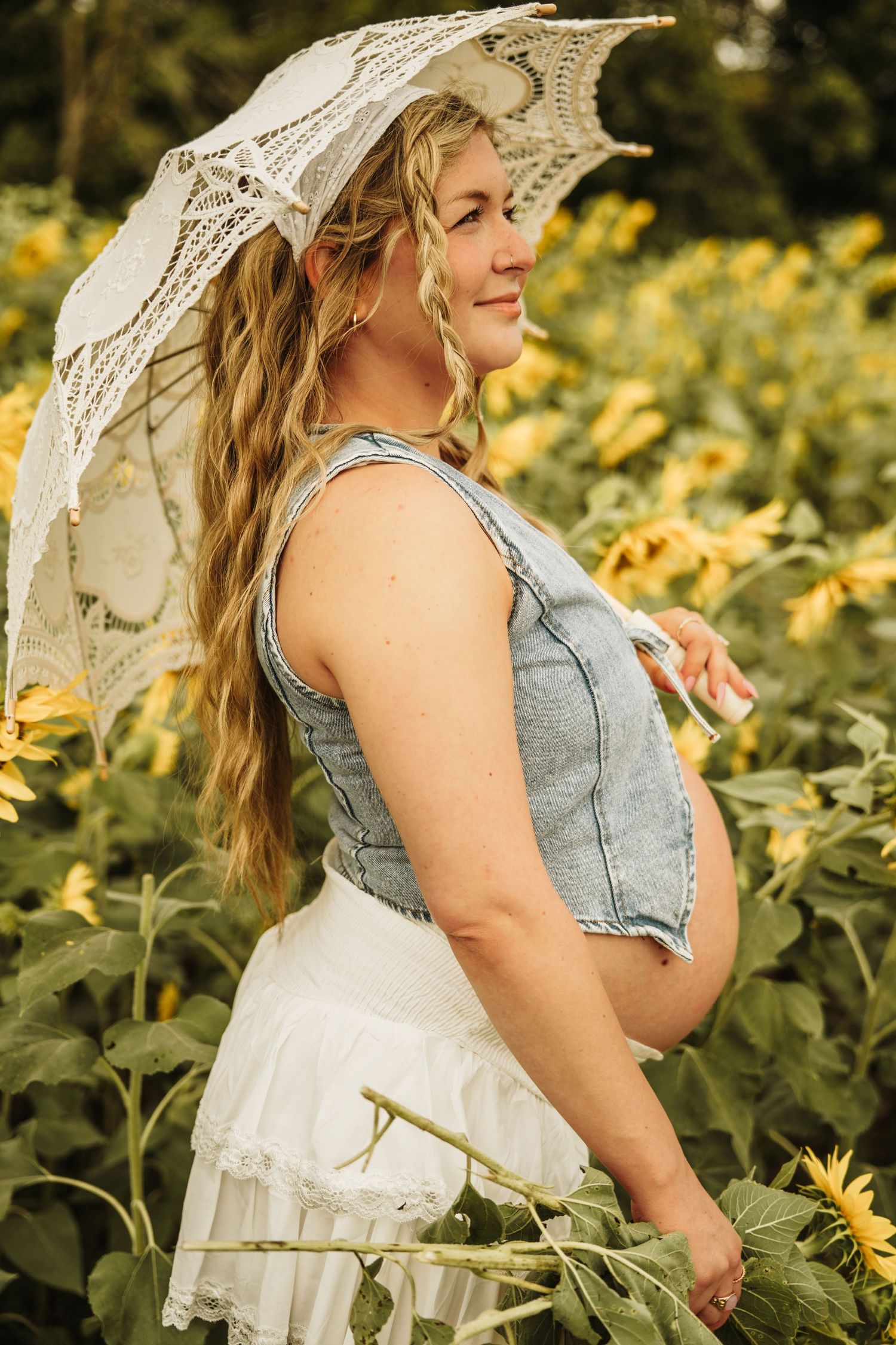 Pregnant woman in denim top and white skirt holding lace umbrella in sunflower field.