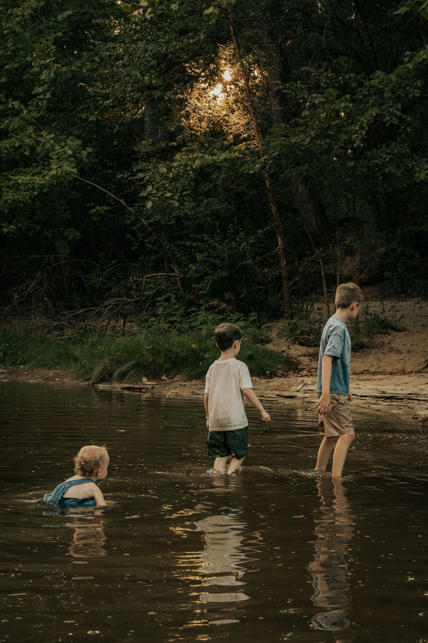 Three children playing in a shallow forest river at dusk, with golden sunlight filtering through green trees.