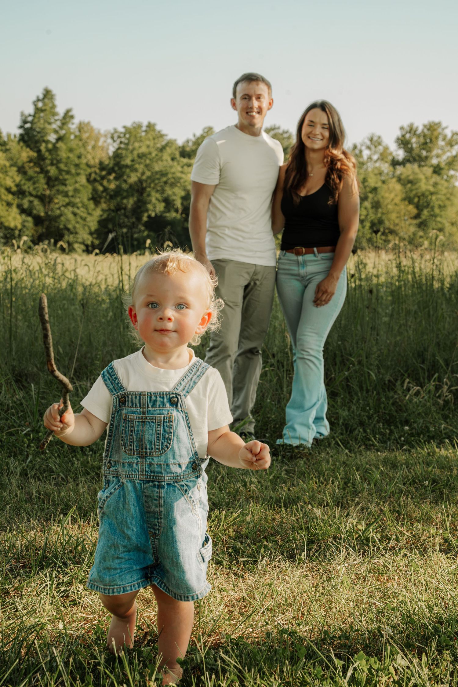 Young family with toddler in denim overalls holding a stick in a sunny green meadow during golden hour.