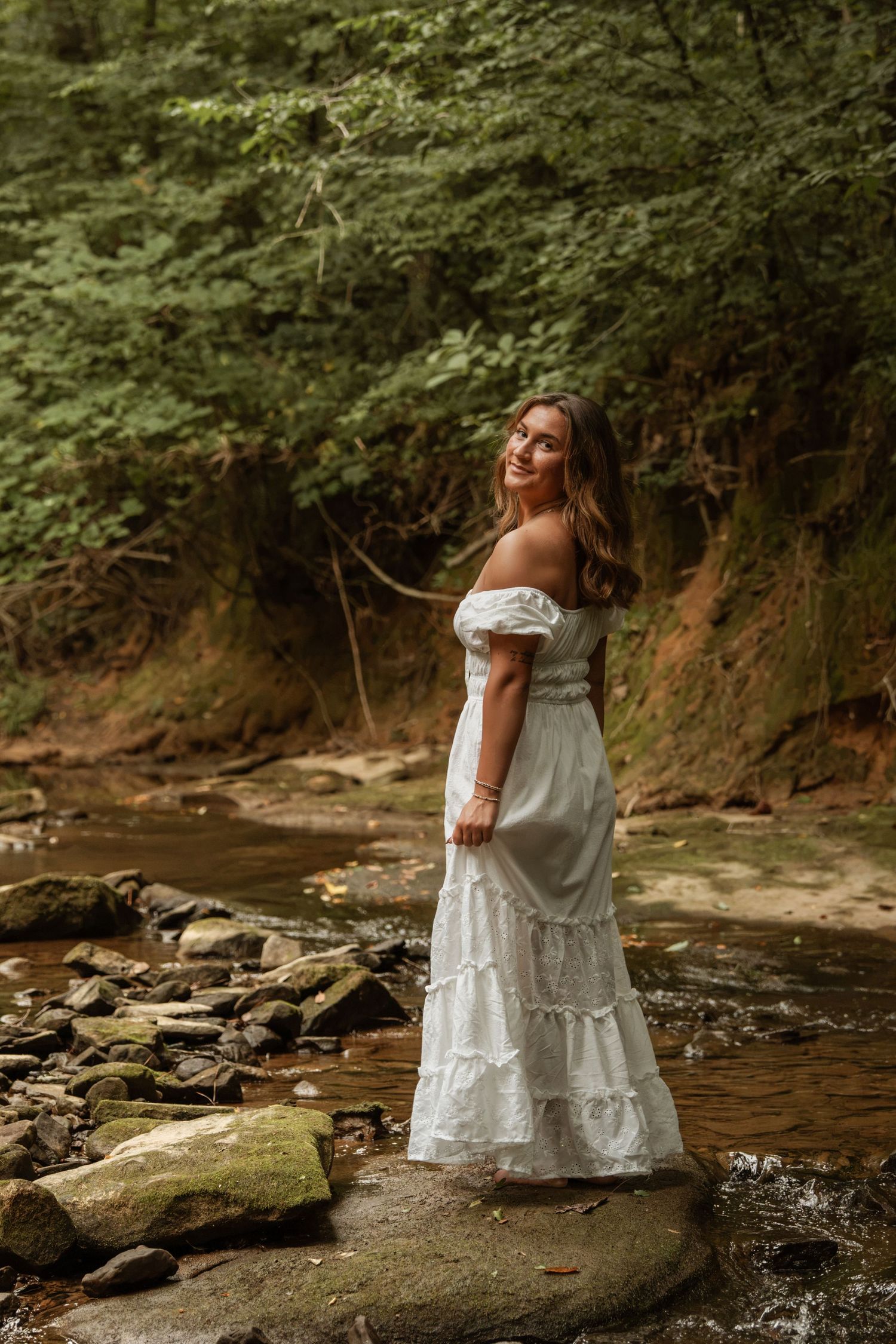Woman in white off-shoulder maxi dress standing on mossy rocks by a forest stream surrounded by lush green trees.