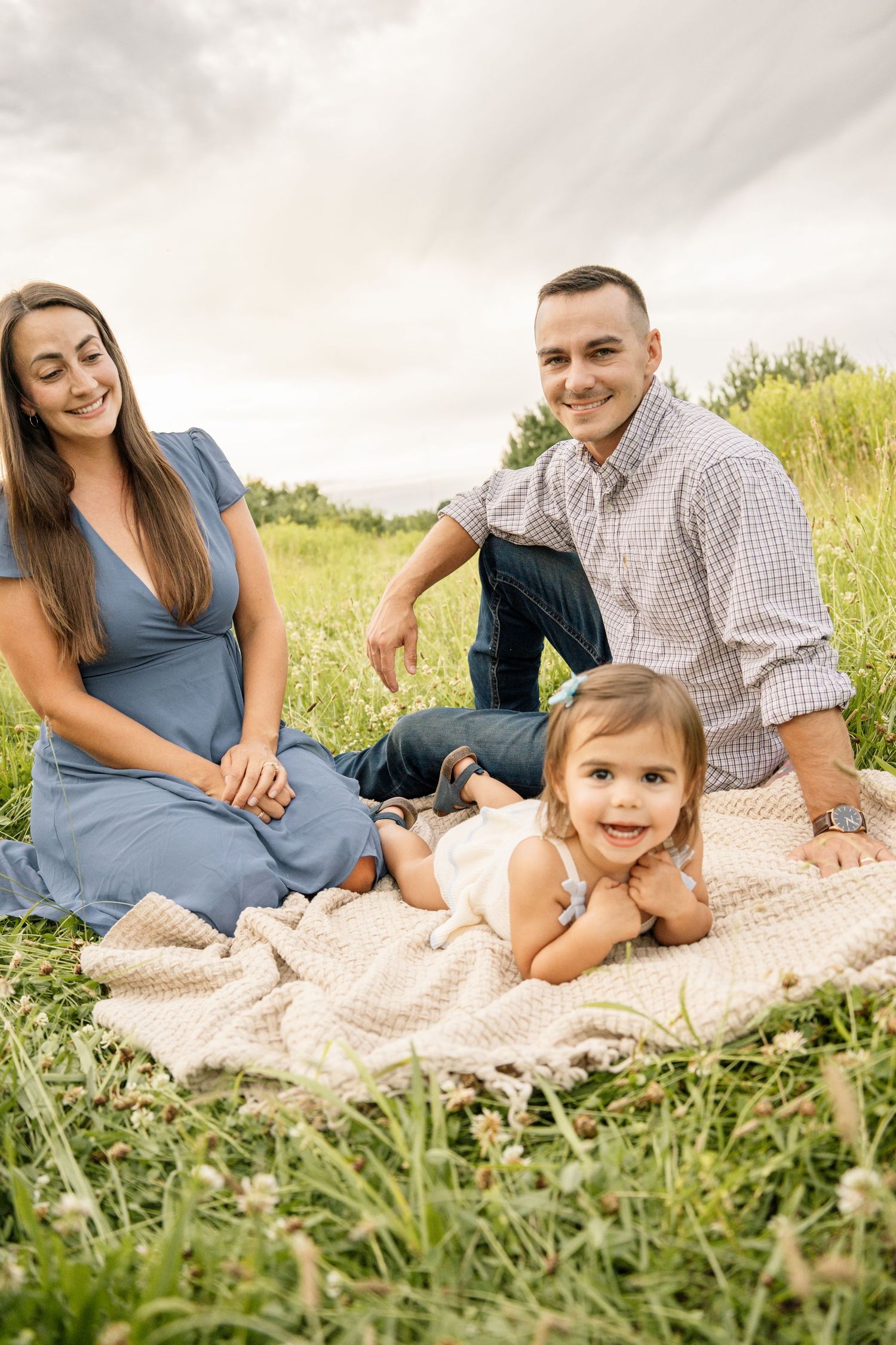 Happy family of three sitting on a blanket in a green meadow, with toddler lying down smiling at the camera.
