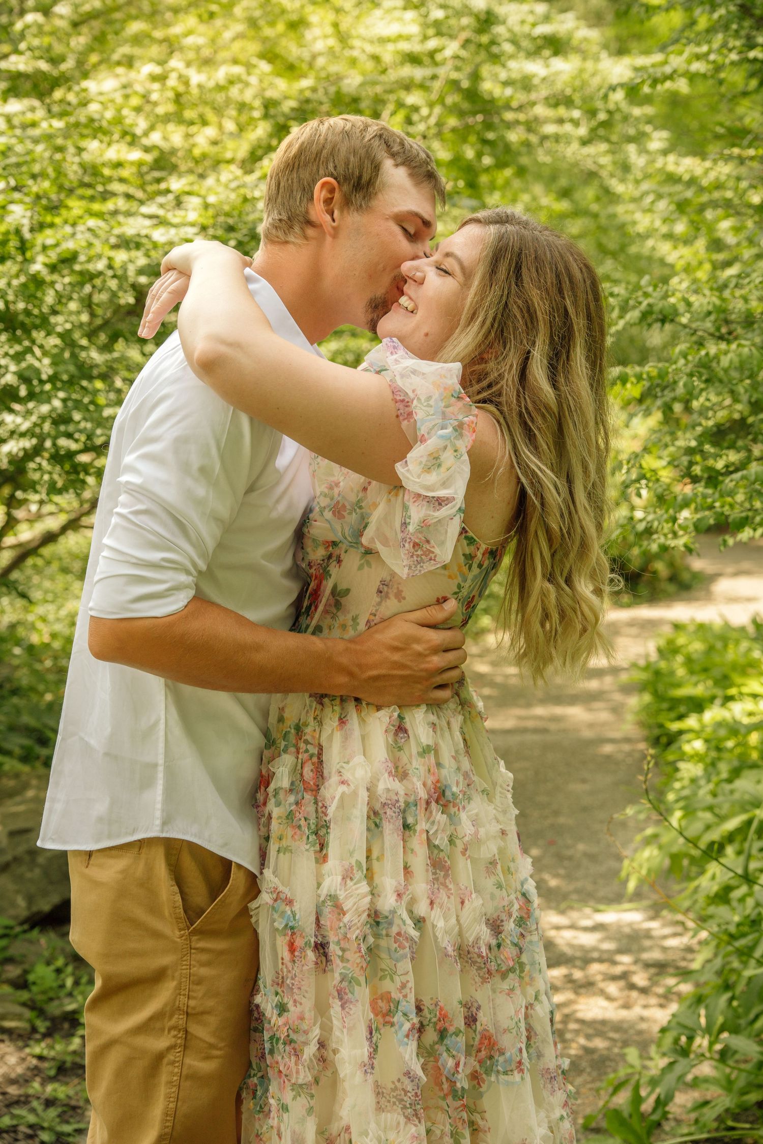 Couple embracing and kissing in a sunlit garden path surrounded by vibrant green foliage.