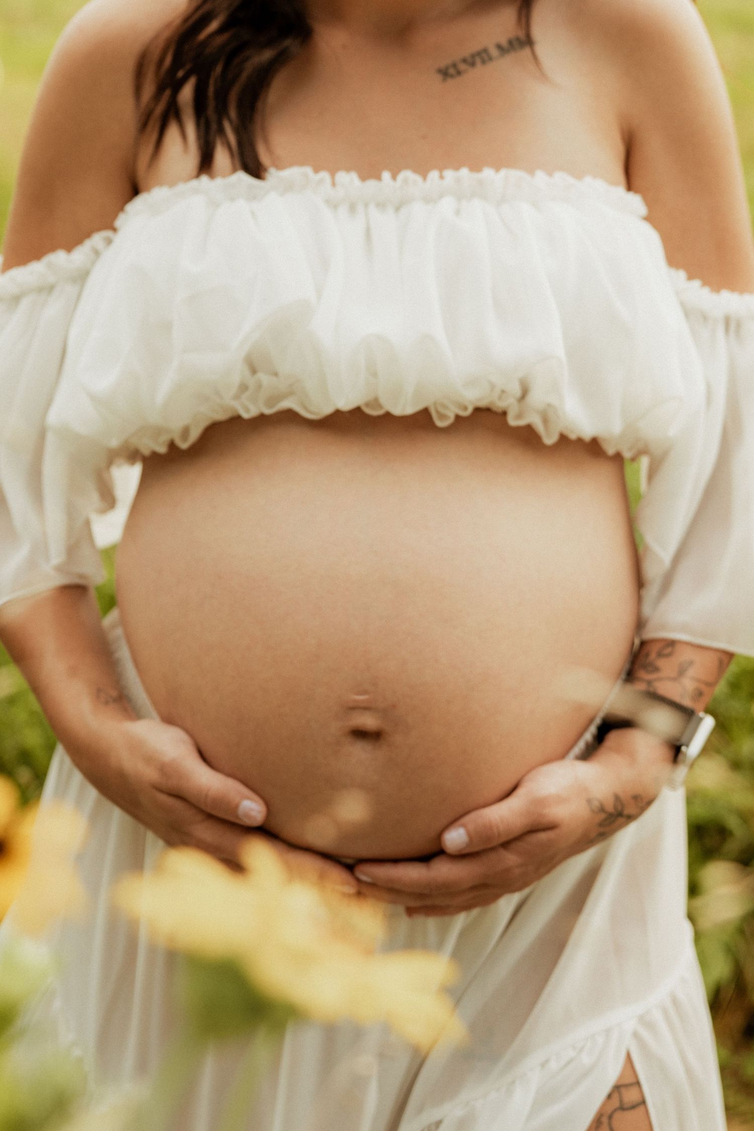 Pregnant woman in white off-shoulder dress cradling her baby bump outdoors among sunflowers.