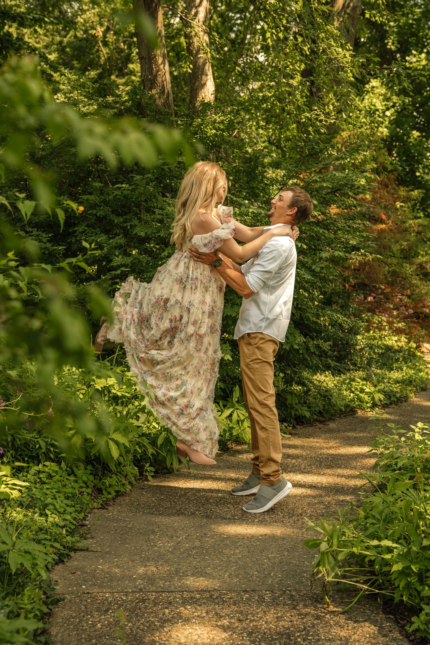 A couple dances joyfully on a garden path surrounded by lush green trees and shrubs on a sunny day.