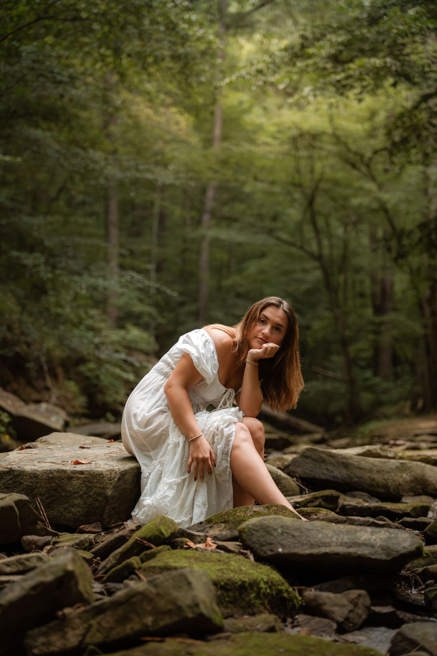 A young woman in a white dress sits gracefully on mossy rocks in a lush green forest setting.