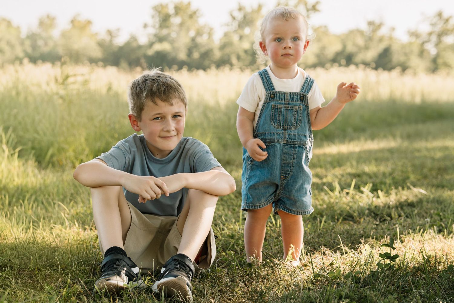 Two young boys pose outdoors in a sunny green field, one sitting cross-legged and one standing in denim overalls.