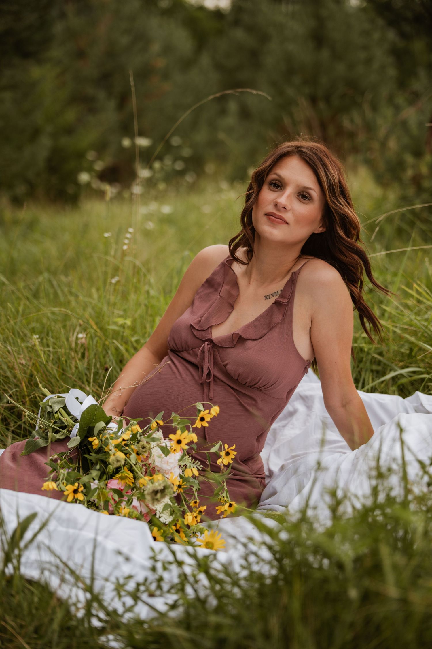 Pregnant woman in mauve slip dress sits on white blanket in tall grass holding wildflower bouquet.